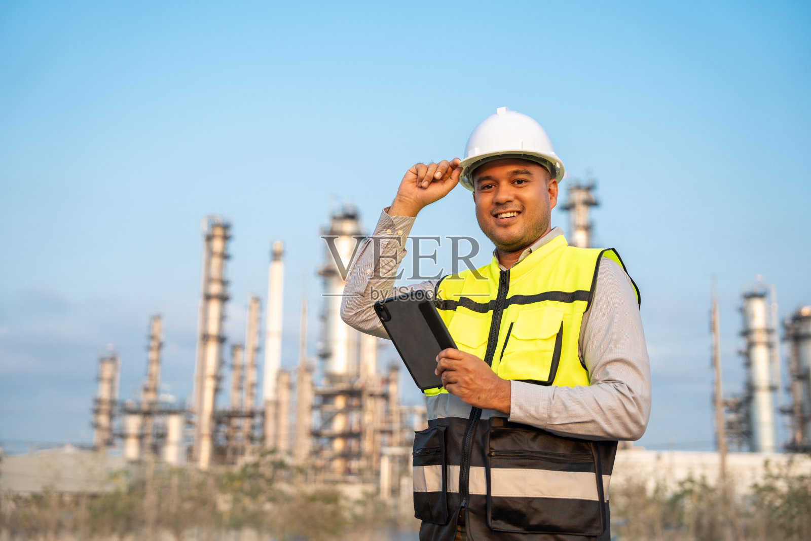 戴着安全帽的亚洲工程师站在炼油厂前。工业园区天然气石化。工厂储油罐和管道。工人们在炼油厂建筑大楼里工作照片摄影图片