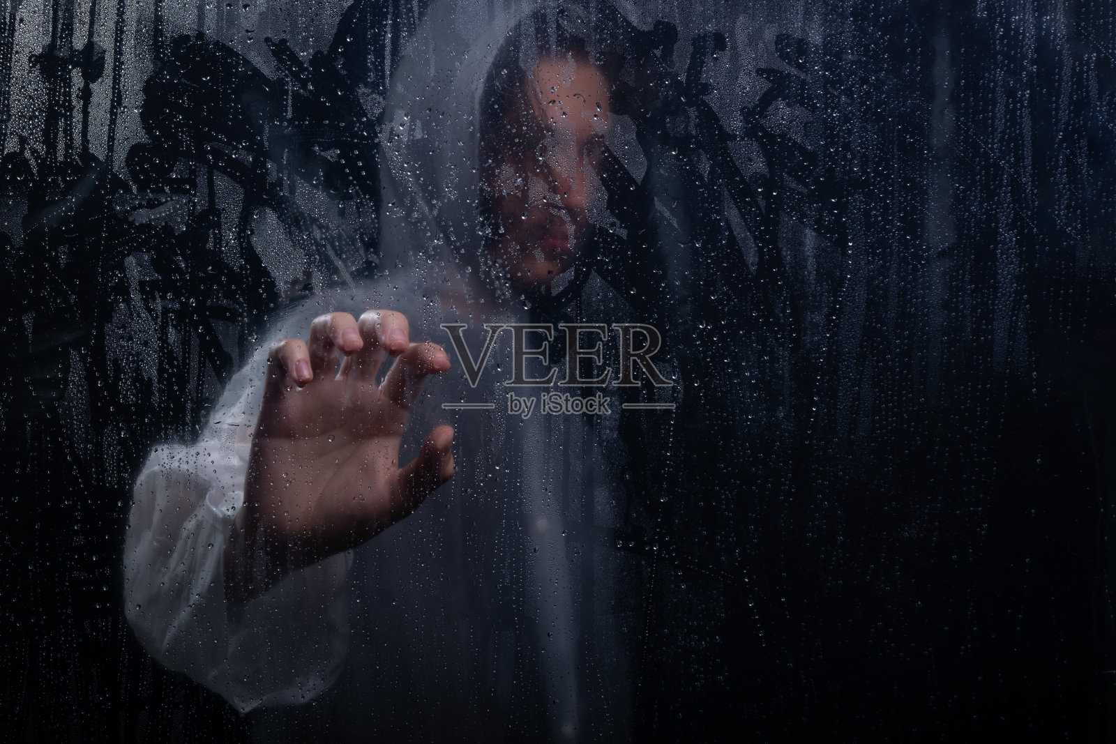 在湿玻璃后面穿着雨衣的人的画像。用手触摸杯子。工作室肖像。照片摄影图片