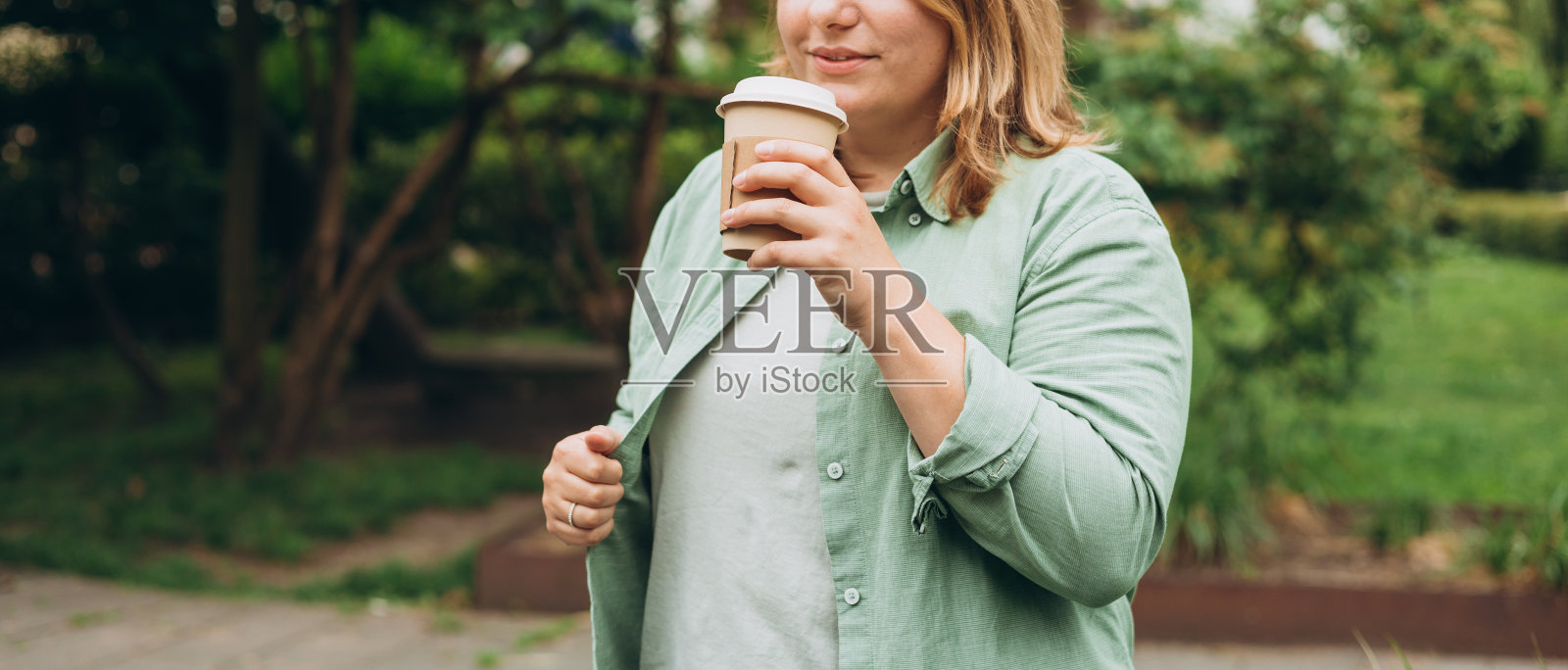 快乐的红发30多岁的女人拿着棕色纸杯咖啡在公园的背景下，咖啡在城市的街道上。吃饭，休息，带走的概念。照片摄影图片