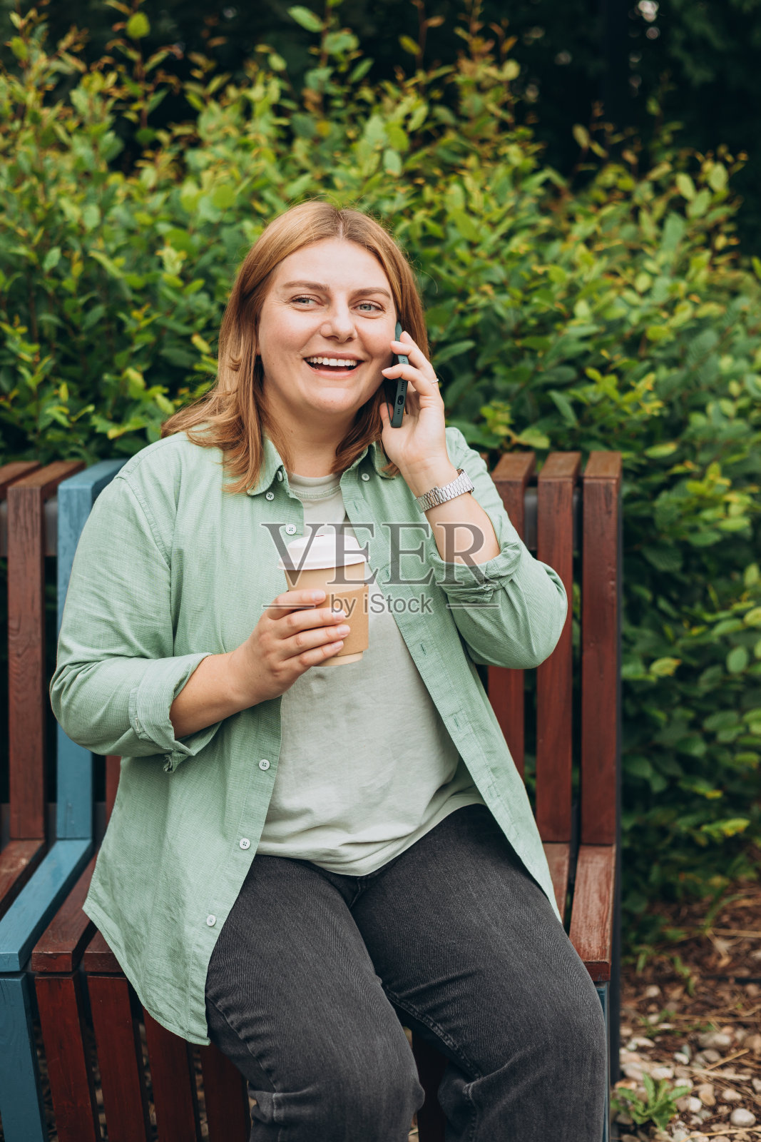 快乐的红发女人穿着时尚的衣服和生态茶在电话里聊天。模型。美丽时尚的女孩打电话智能手机，坐在户外的长凳上，拿着咖啡杯带走。照片摄影图片
