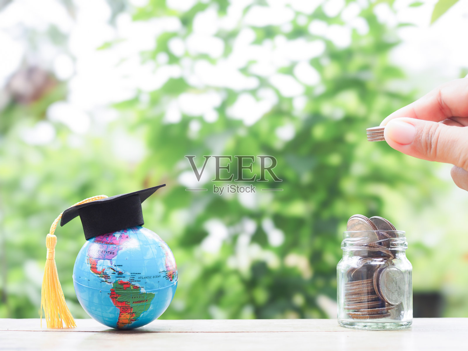 近景:将硬币放入玻璃瓶的手和戴着毕业帽的地球仪。为教育存钱的观念，学生贷款，奖学金，未来的学费照片摄影图片