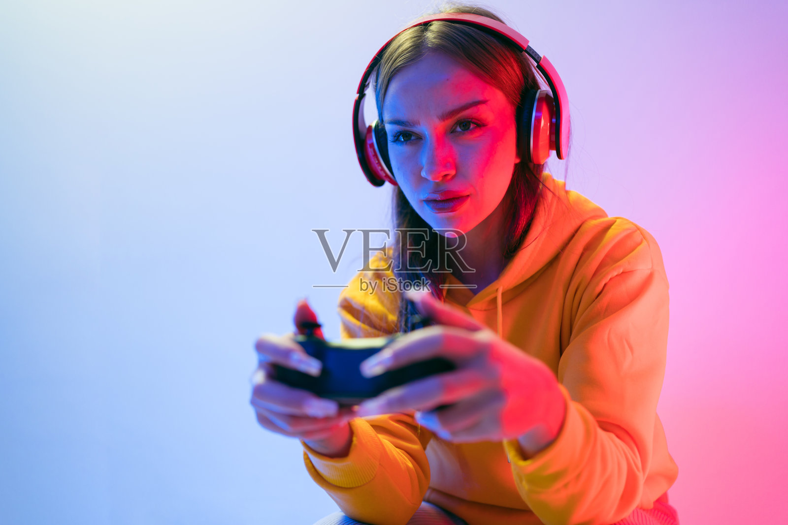 一个年轻的白人美女穿着黄色连帽衫，用操纵杆和耳机玩着电子游戏，玩得很开心，专注，惊喜，还有一张蓝色和红色霓虹灯的胜利脸。照片摄影图片