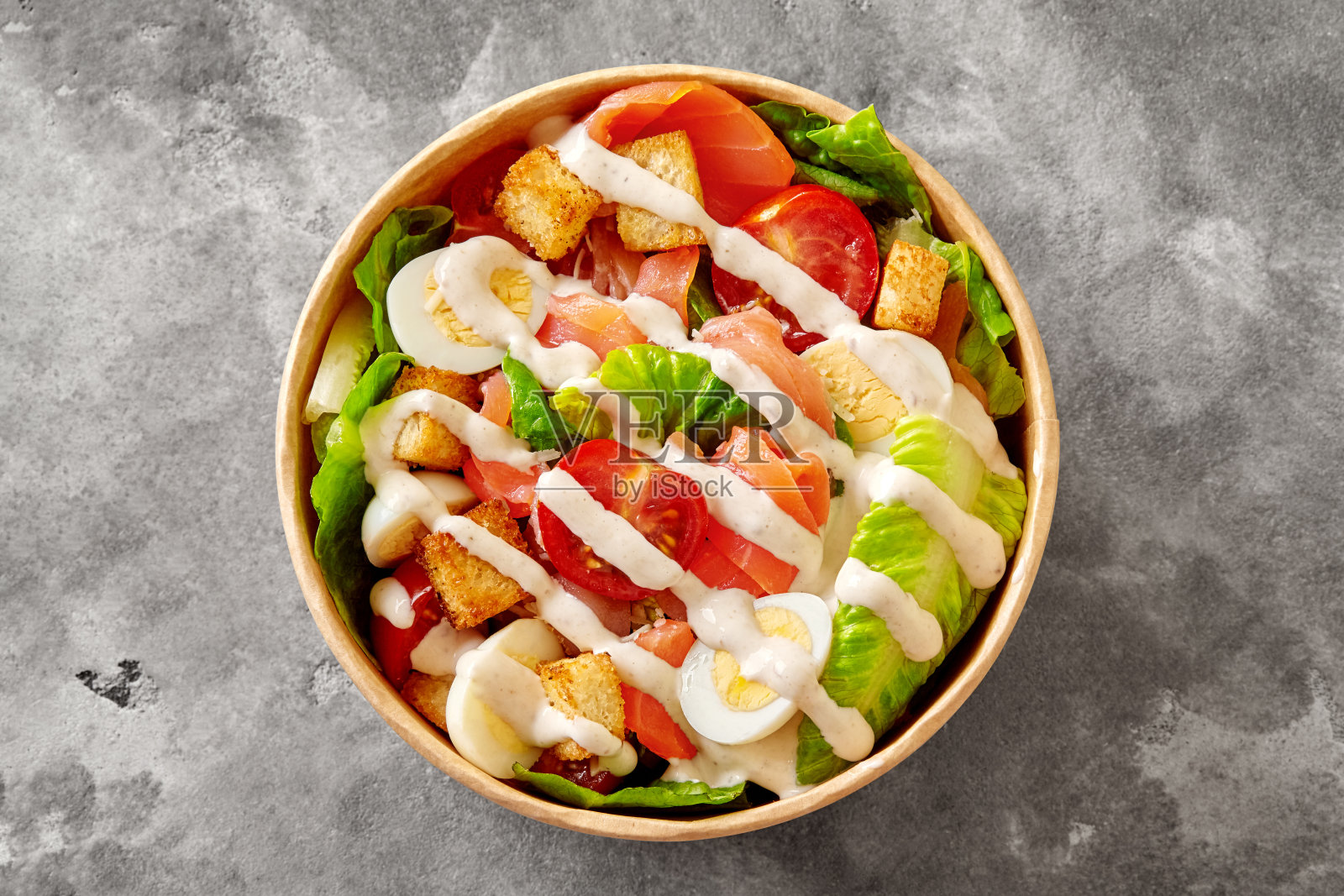 纸碗里有生菜、西红柿、鹌鹑蛋、面包丁和烟熏鲑鱼的凯撒沙拉，表面是灰色的石头照片摄影图片