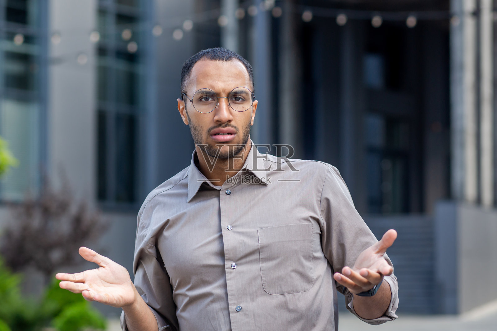戴眼镜站在办公楼外的年轻西班牙裔男子特写肖像，担忧地看着镜头，张开双臂照片摄影图片