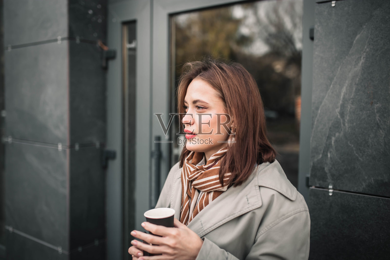 美丽的年轻女子在城市里端着一杯咖啡穿着大衣，戴着围巾。照片摄影图片