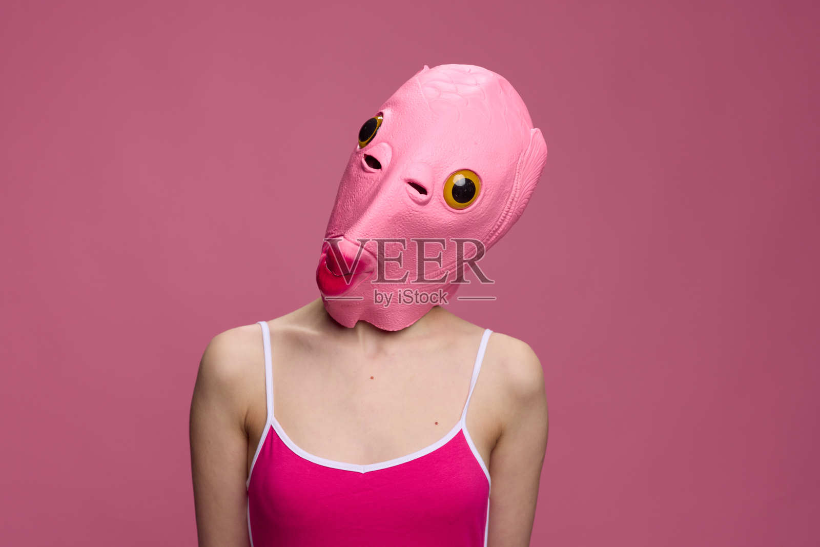 一个戴着粉红色鱼头万圣节面具的女人站在那里，看着粉红色背景下的相机照片摄影图片