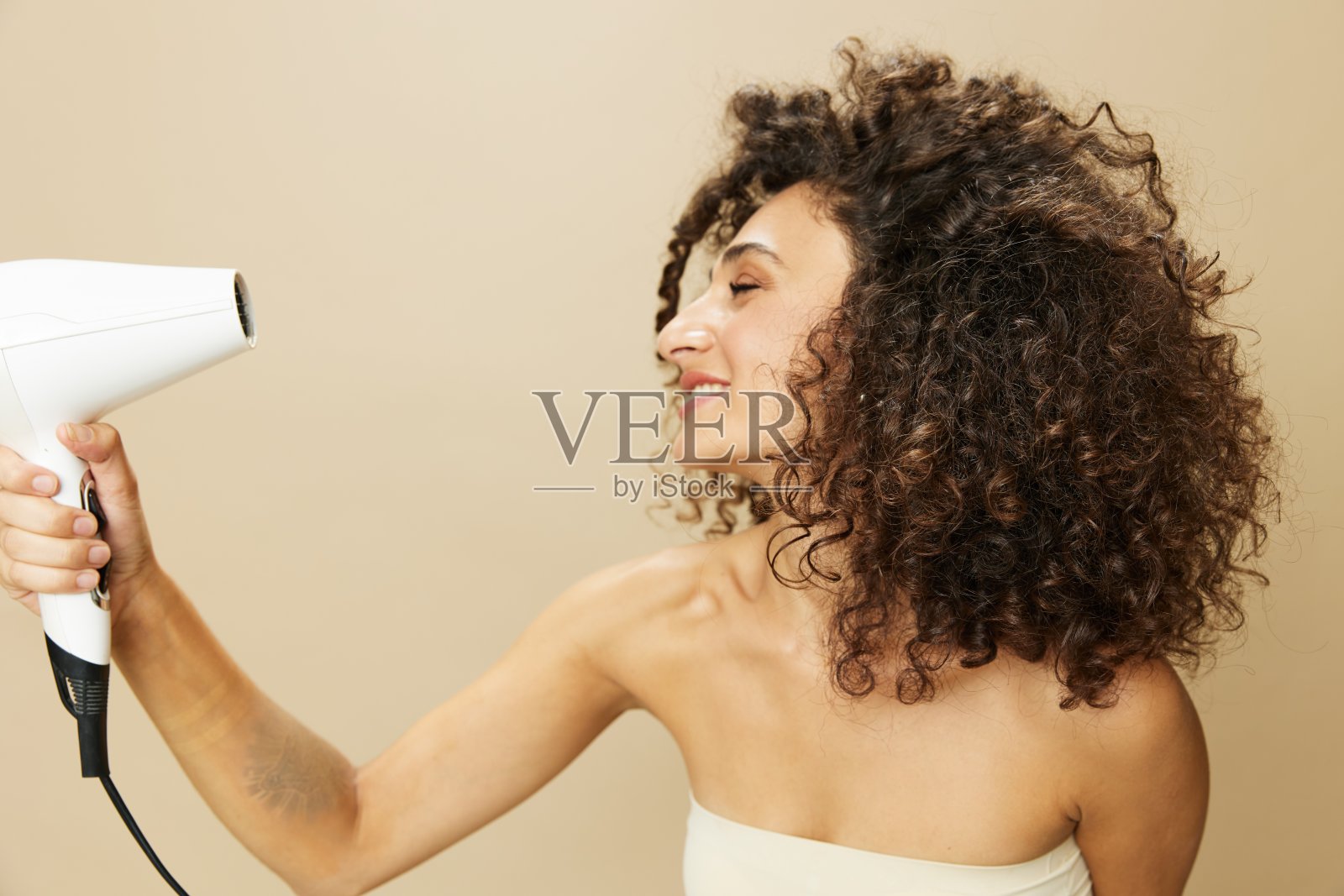 女人用吹风机吹干卷曲的非洲发型，家用美容护理定型产品，米色背景上的微笑照片摄影图片