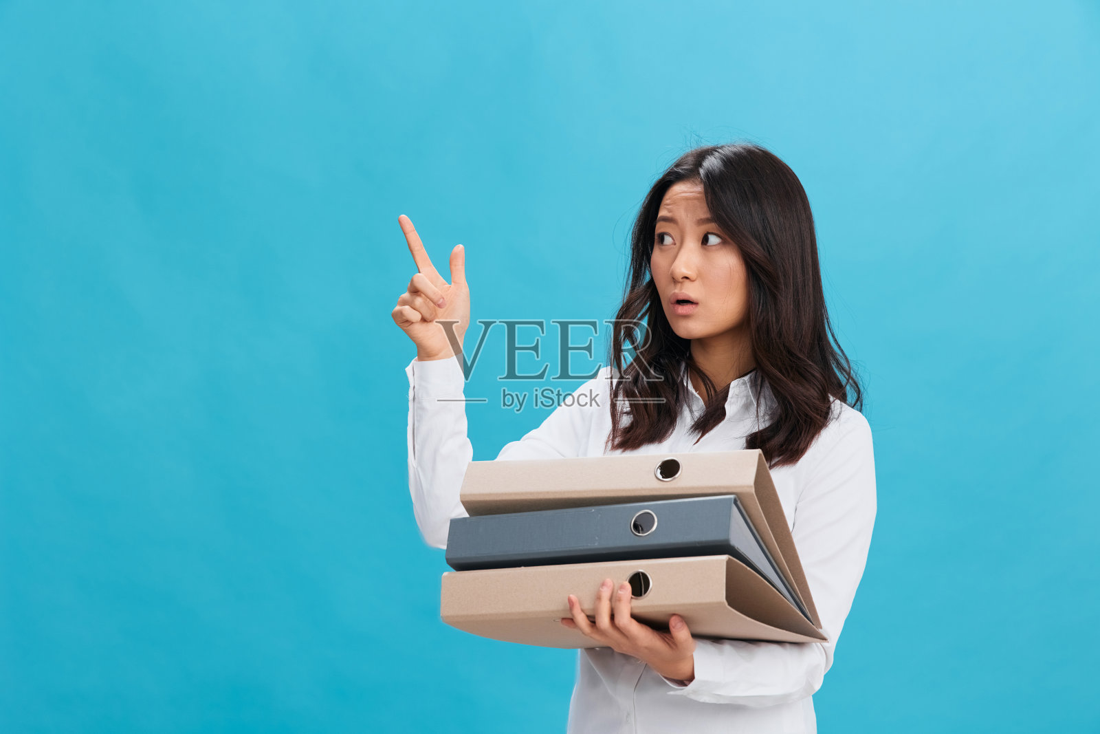 兴奋，震惊，可爱的亚洲女商人，穿着经典的办公室着装，拿着很多文件夹，手指向上，在蓝色的工作室背景上孤独地摆姿势。很酷的生意机会。太多的工作任务概念照片摄影图片