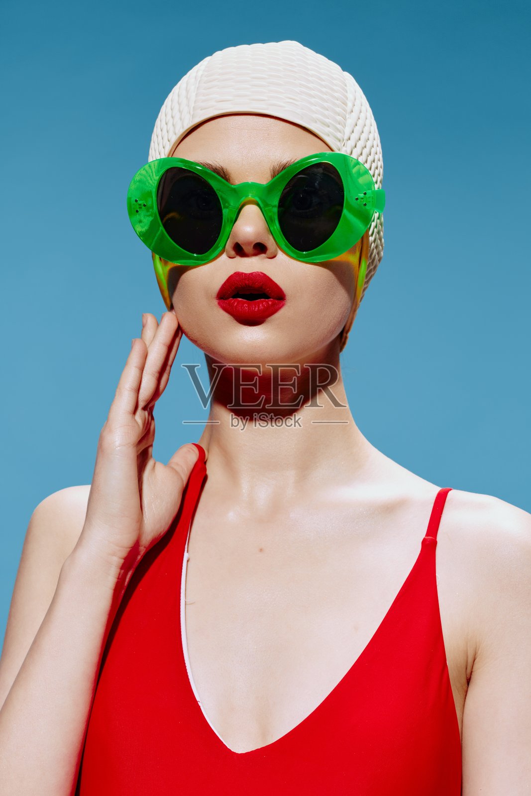 在蓝色背景的摄影棚里，一个戴着游泳帽、穿着红色泳衣的震惊的女女孩张开了她的嘴。艺术Nft时尚理念照片摄影图片