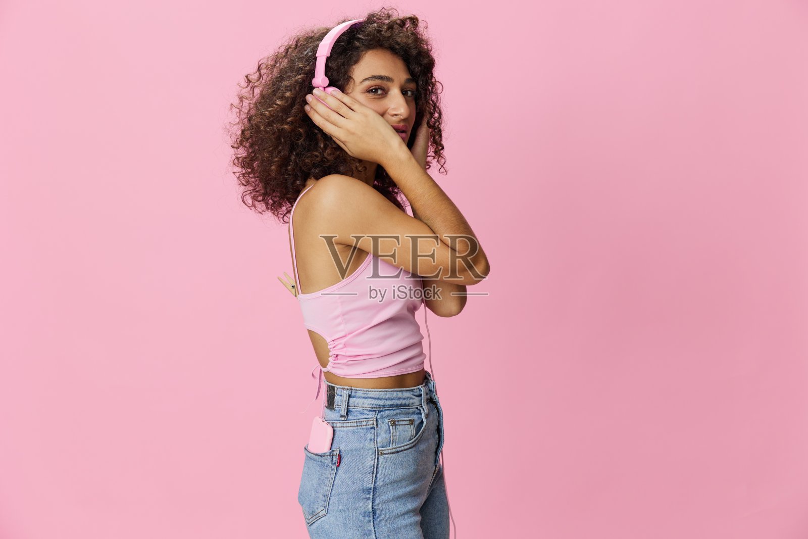 快乐的女人戴着耳机，卷发，闭着眼睛听着音乐和唱歌，穿着粉红色的t恤和牛仔裤，在粉红色的背景DJ派对上，复制空间照片摄影图片