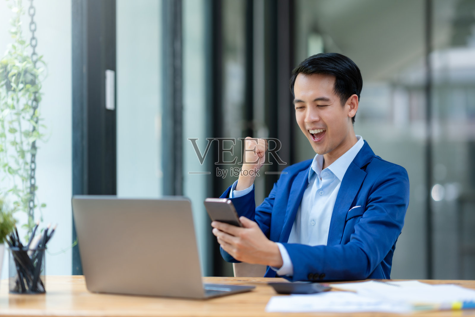 成功的亚洲商人从手机中收到好消息。快乐的亚洲商人举起胜利的手，用智能手机开心地微笑。工作中成功的概念。照片摄影图片