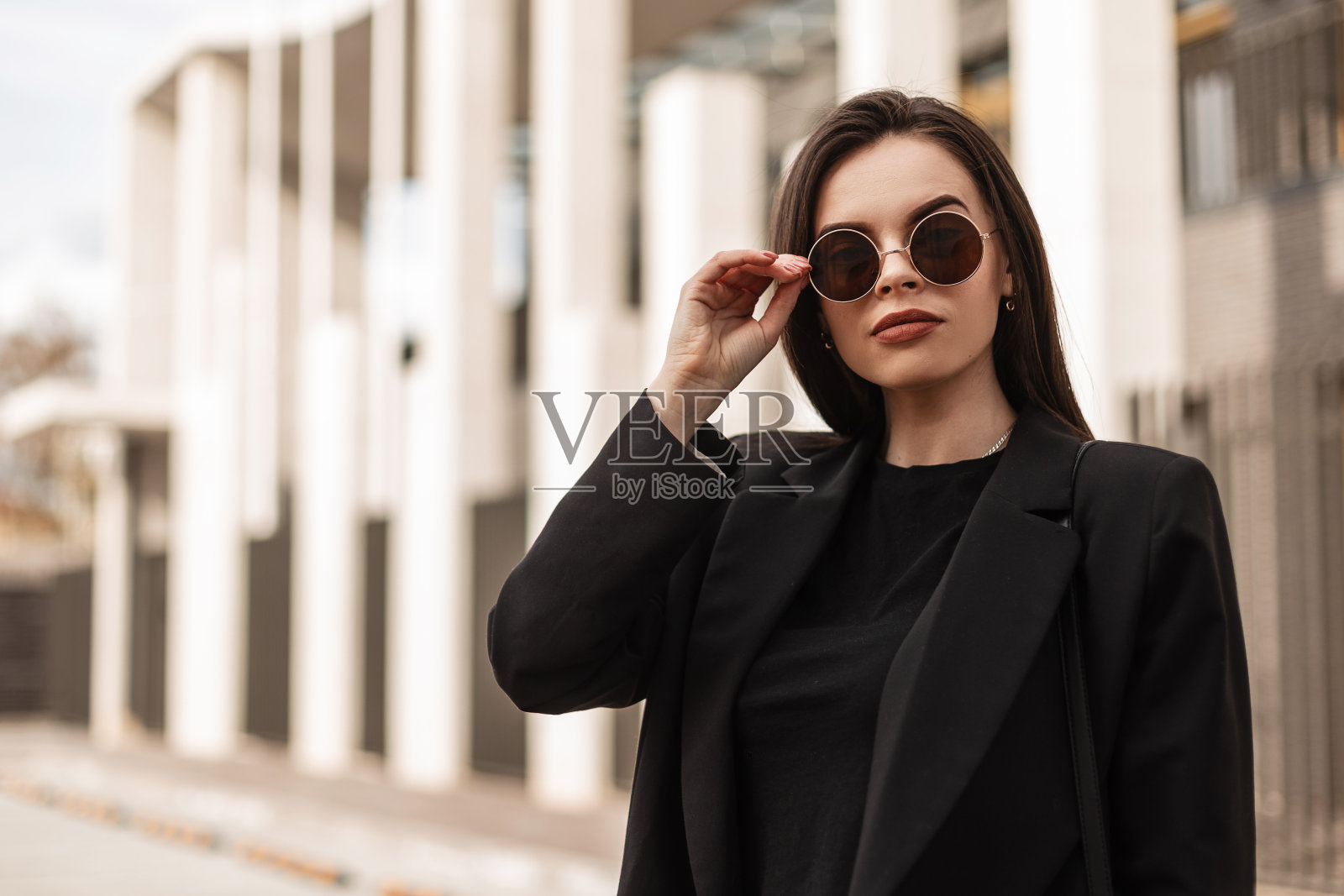 肖像华丽时髦的年轻女子在时尚的青年黑色夹克的时尚太阳镜附近的白色复古墙户外。可爱的女孩模特穿着时尚的运动上衣，在街上矫正圆眼镜。照片摄影图片