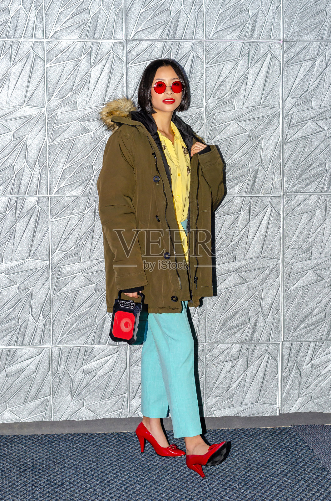 美丽的年轻女子在工作室里穿着五颜六色的衣服，一件卫衣夹克，戴着红色太阳镜，拿着一个复古的音响扬声器，背景是现代的墙照片摄影图片