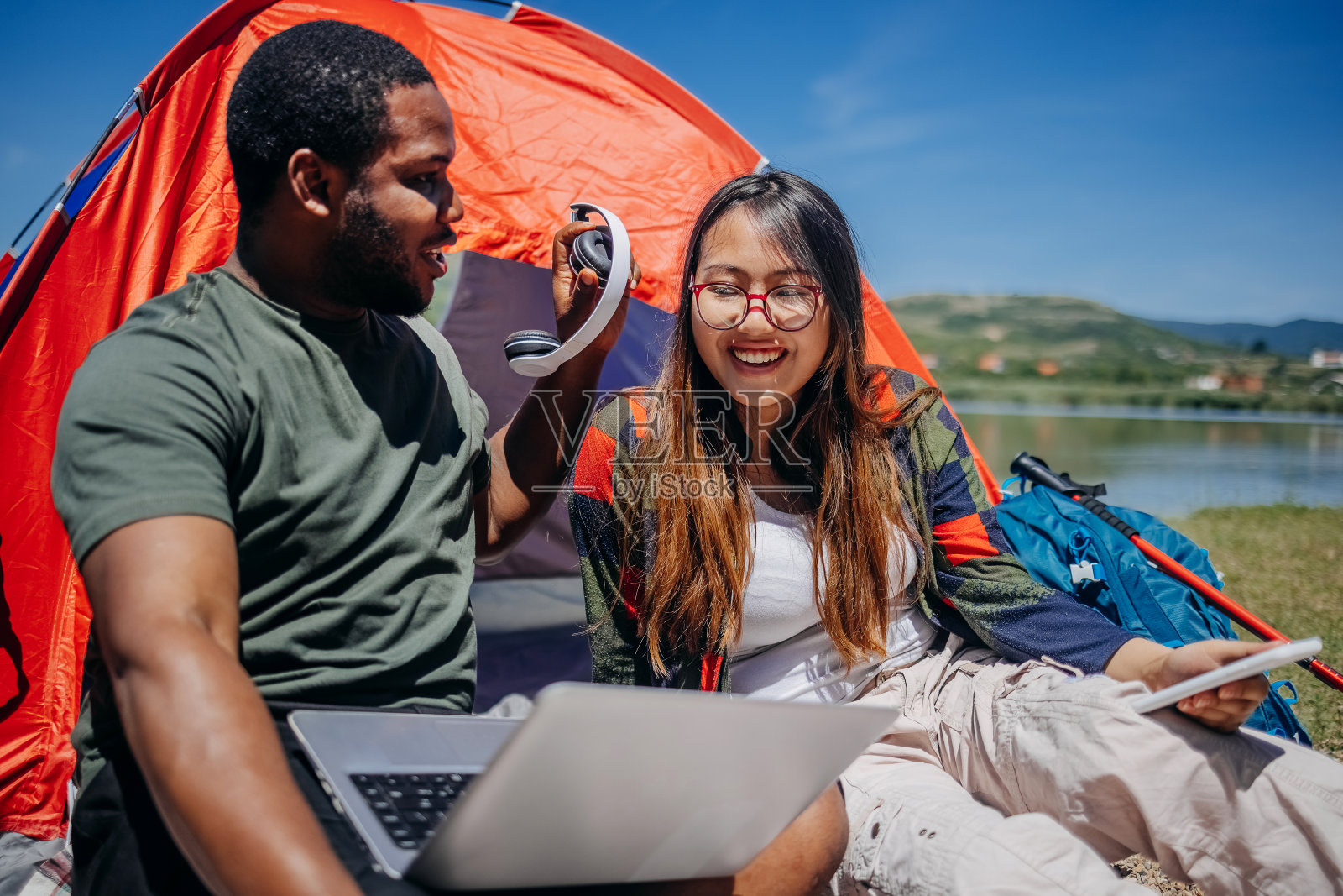 年轻的多种族夫妇一起在湖边露营，坐在帐篷前，用无线耳机听音乐照片摄影图片