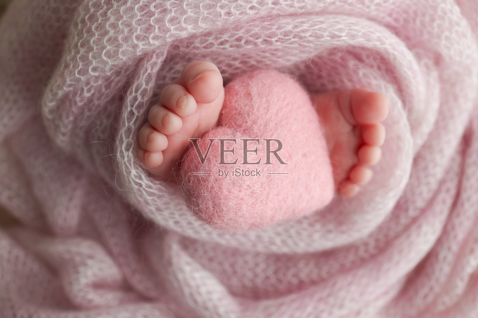 新生婴儿的小脚。新生儿柔软的脚裹在粉红色的羊毛毯子里。新生儿脚趾、脚跟和脚的特写。在婴儿的腿上编织粉红色的心。微距工作室摄影。照片摄影图片
