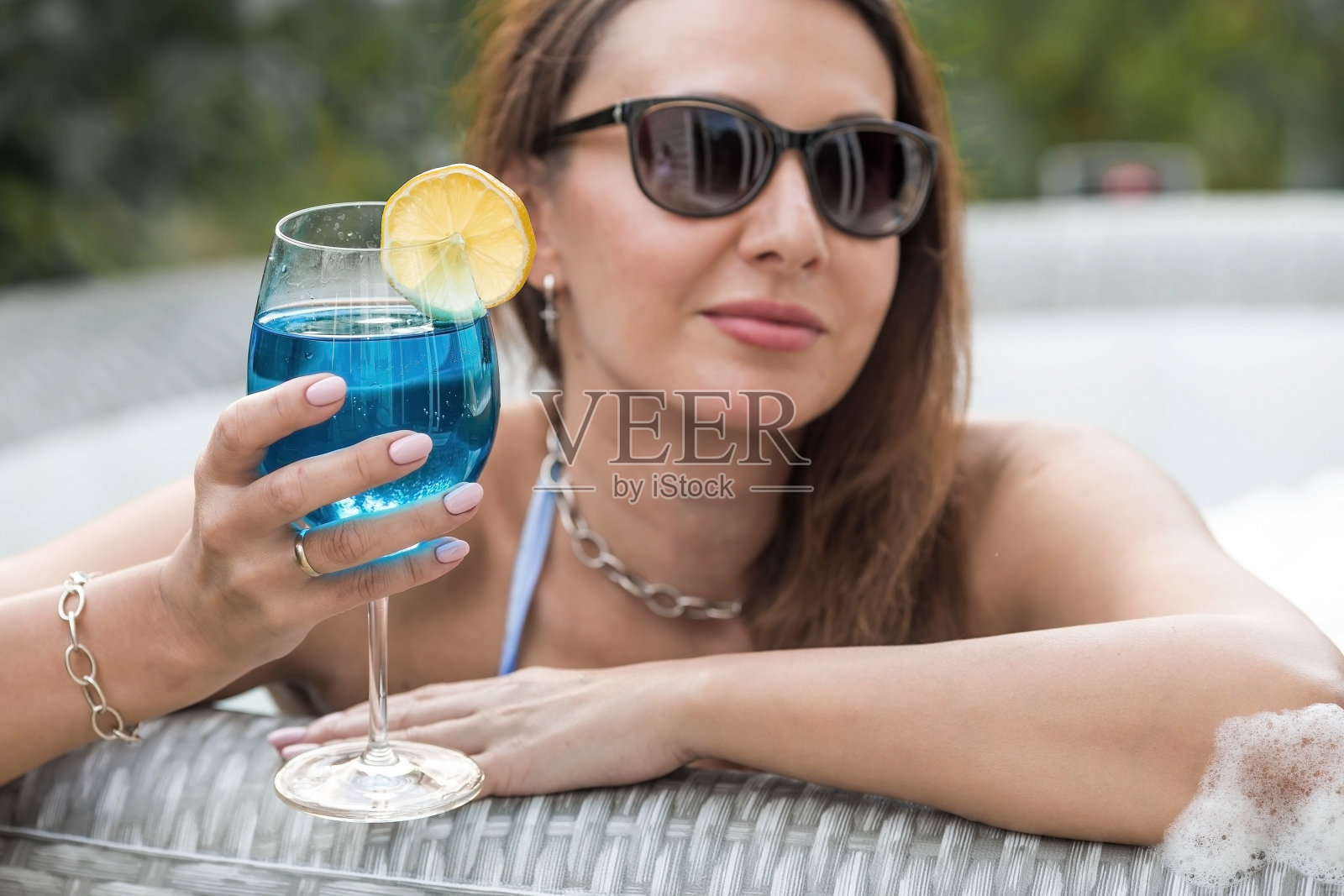 放松的概念，水疗和泡沫派对。一个穿着泳衣、戴着太阳镜的年轻女子坐在按摩浴缸里，一杯蓝色的泻湖鸡尾酒和一颗黄色的柠檬在泳池的背景上。照片摄影图片