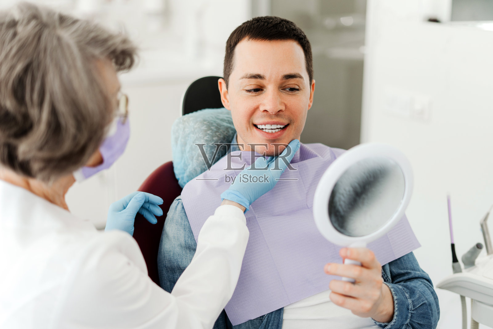 微笑迷人的男病人拿着镜子，看着，和资深专业牙医交谈照片摄影图片