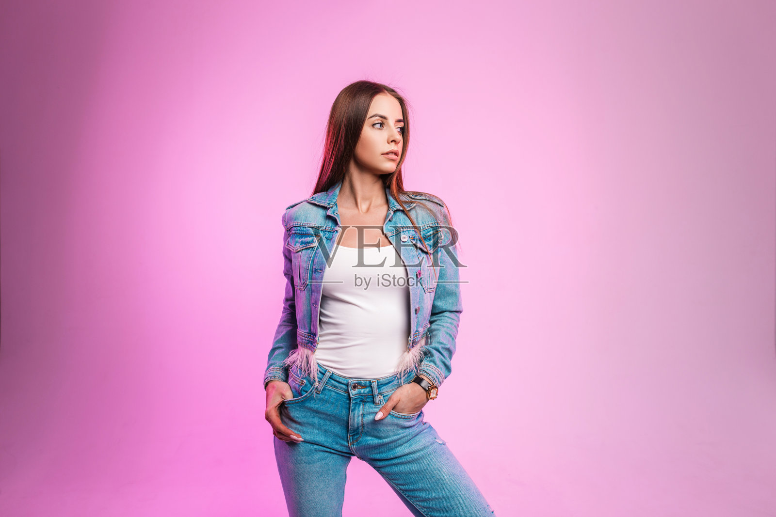 现代迷人的年轻女子穿着蓝色时尚牛仔夹克，穿着时尚牛仔裤和白色t恤，站在工作室的粉红色墙壁旁。美丽的时尚女孩时装模特在室内摆姿势。休闲风格。照片摄影图片