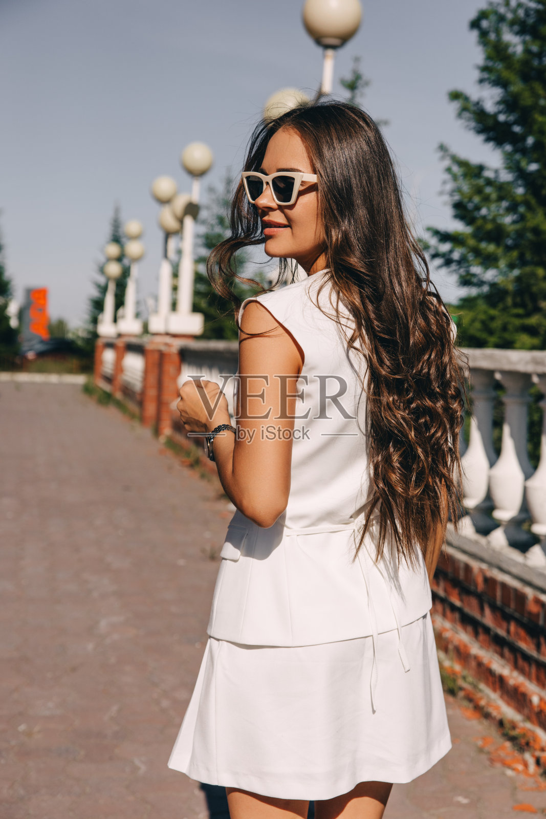 一位身穿白衣、戴着墨镜的年轻女子在一座红砖建筑旁摆姿势散步照片摄影图片