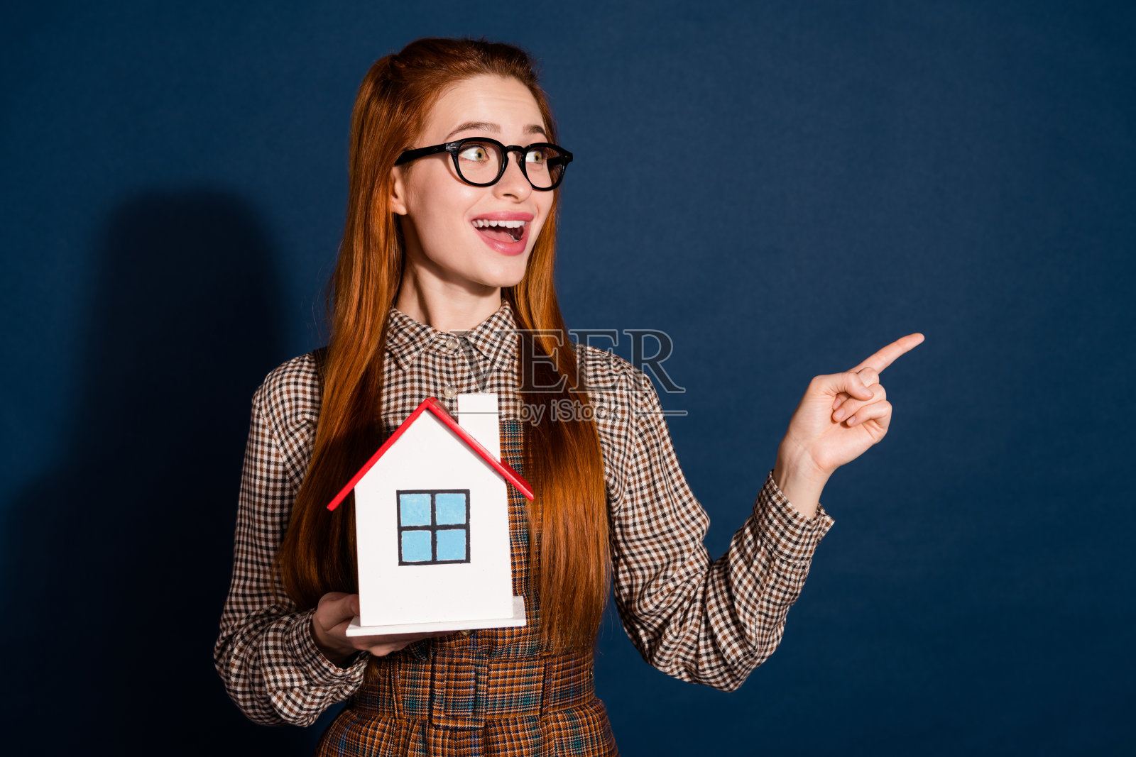 时髦印象深刻的女士穿着格子装抱着房子看着指向空旷的空间孤立的深蓝色背景的照片照片摄影图片
