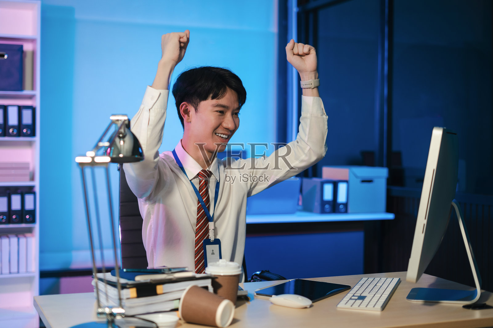 快乐的年轻商人看着成功兴奋的笔记本电脑坐在桌子上举起双手，以“是”的手势庆祝商业成功。照片摄影图片