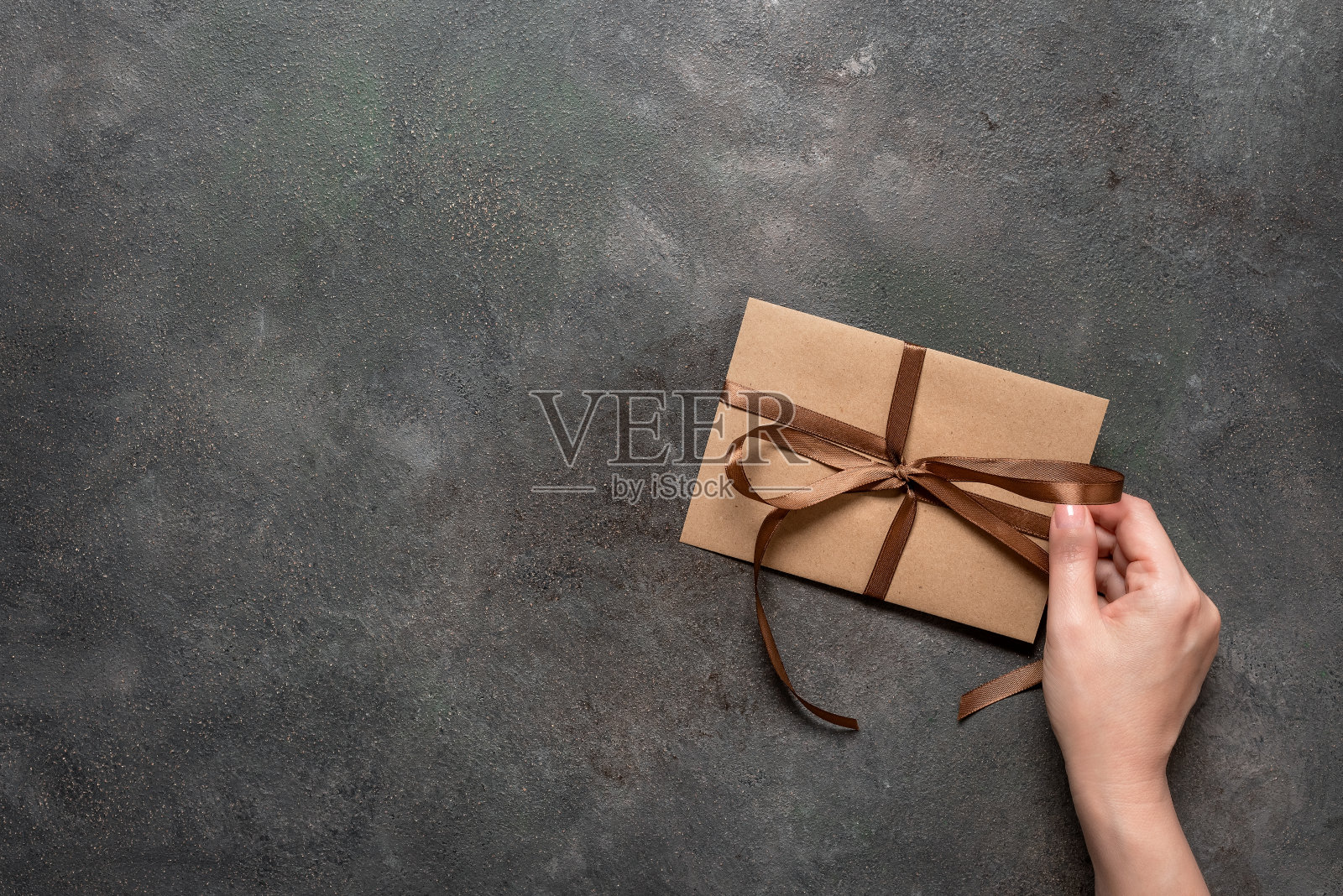 一个女人的手拿着一个用丝带系着的棕色工艺信封，黑色的垃圾背景，俯视图，平躺。副本的空间。假期的邀请。照片摄影图片