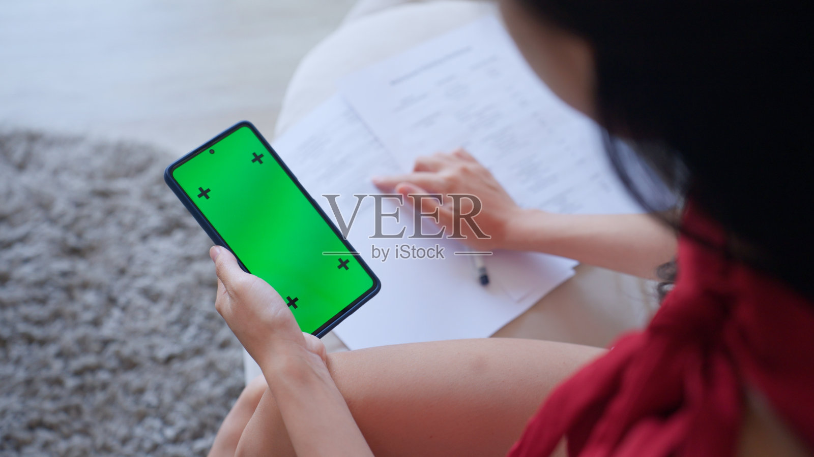 亚洲女性的后视图查看她的个人财务记录在手机上，并在家里写在纸上的费用清单。从事经济金融领域室内工作的年轻成年女性。照片摄影图片