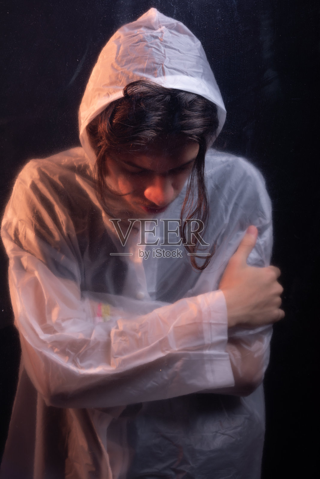 玻璃后面穿着透明塑料雨衣的年轻人。面对隐藏。工作室肖像。照片摄影图片