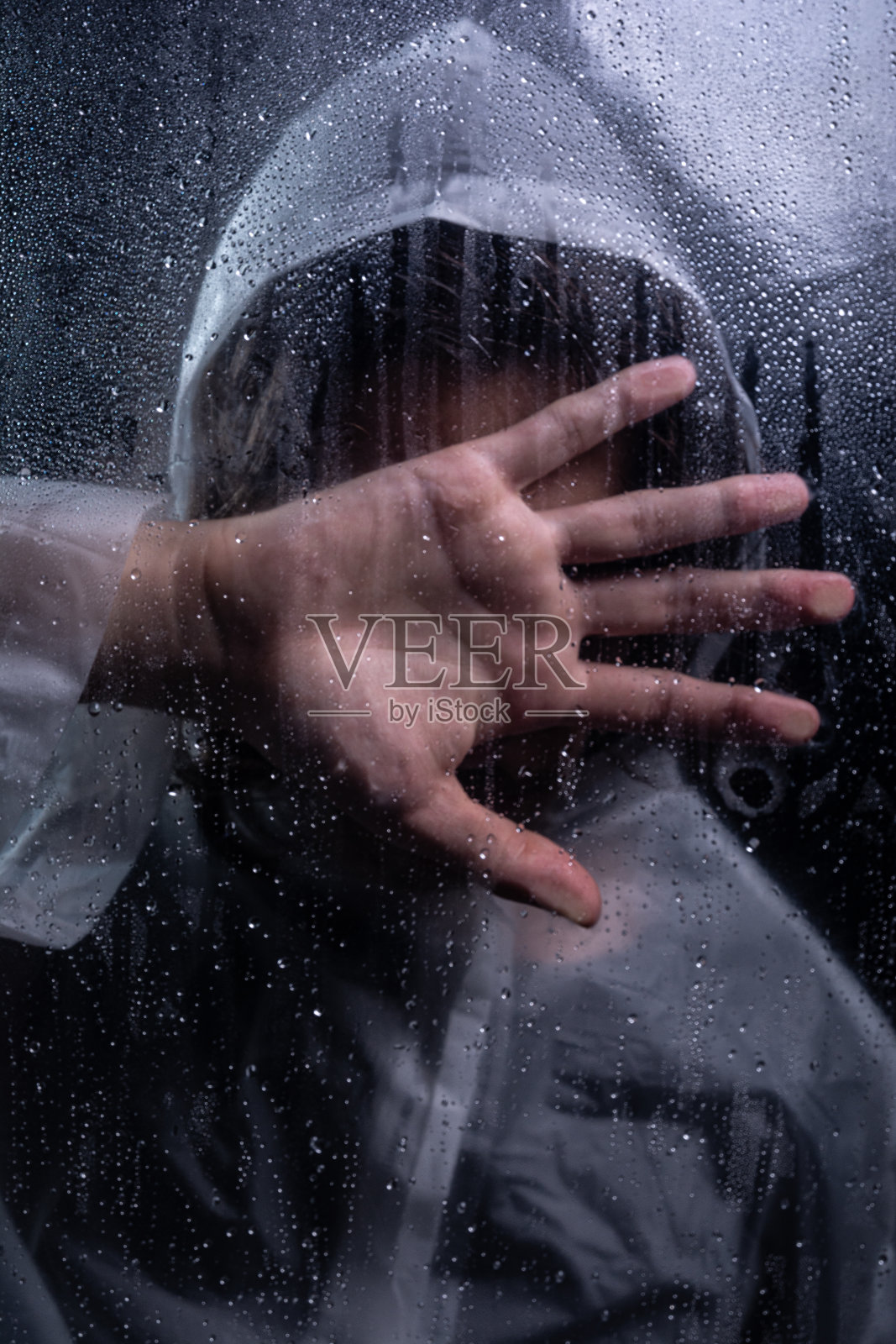 一个男人的特写肖像，他穿着雨衣，手捂着脸，后面是潮湿的玻璃。工作室肖像。照片摄影图片