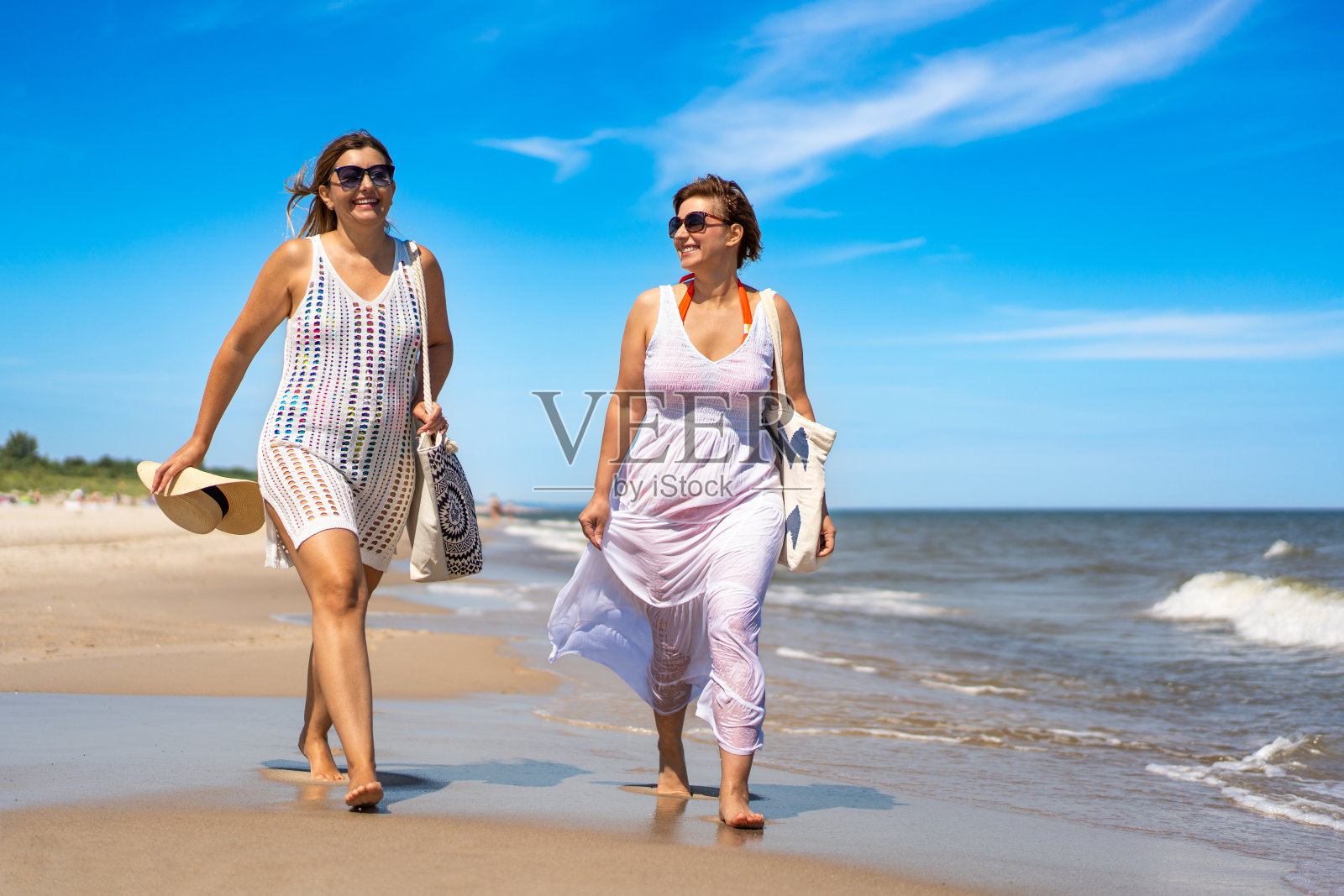 暑假。两个面带微笑的中年妇女，穿着泳衣和白色夏装，提着沙滩袋，边说边从海边走向海滩。漫步在波罗的海的沙滩上。前视图照片摄影图片
