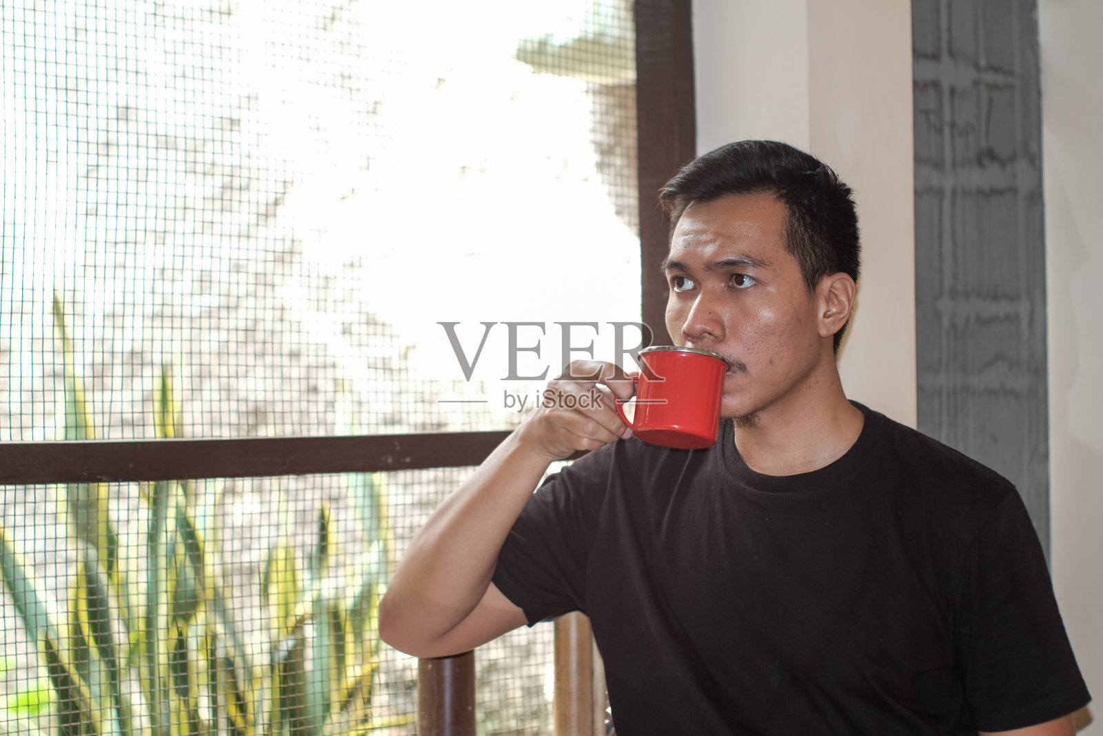 一名东南亚男子的画像，他坐在阳台上的竹椅上，手里拿着红色的铁杯咖啡照片摄影图片