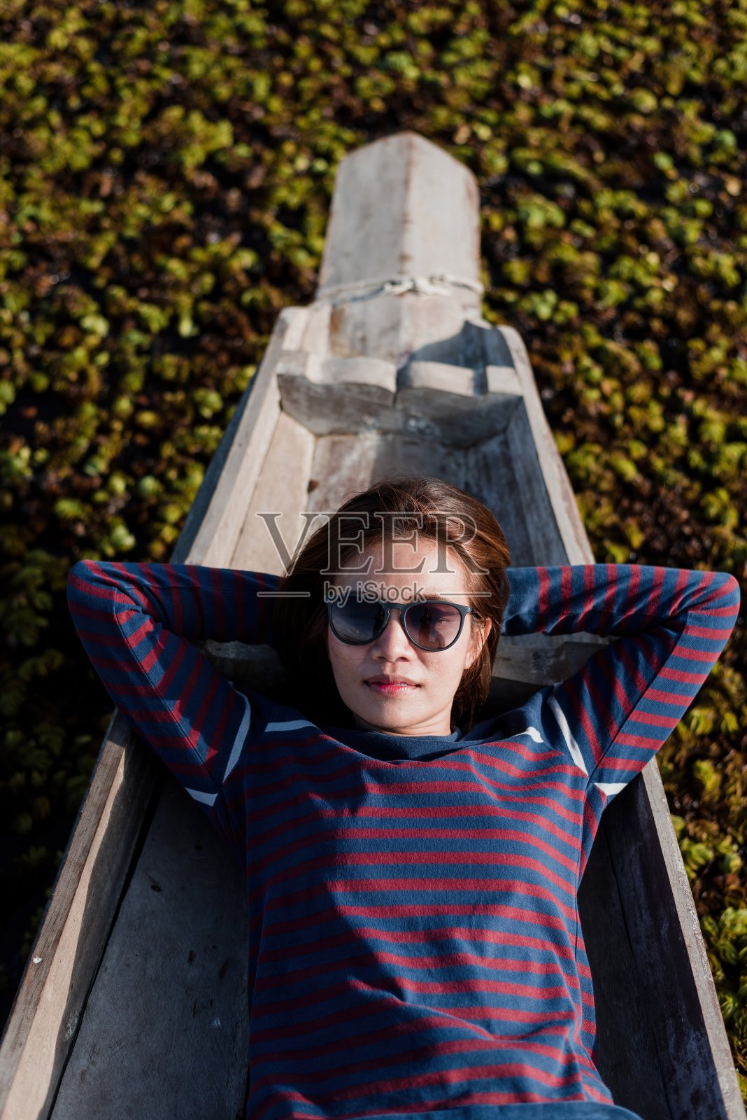 一个年轻女子在湖上的木船上放松，周围是绿色的树叶。她的太阳镜和平静的水面营造出一种宁静的氛围，非常适合暑假或旅行摄影。照片摄影图片