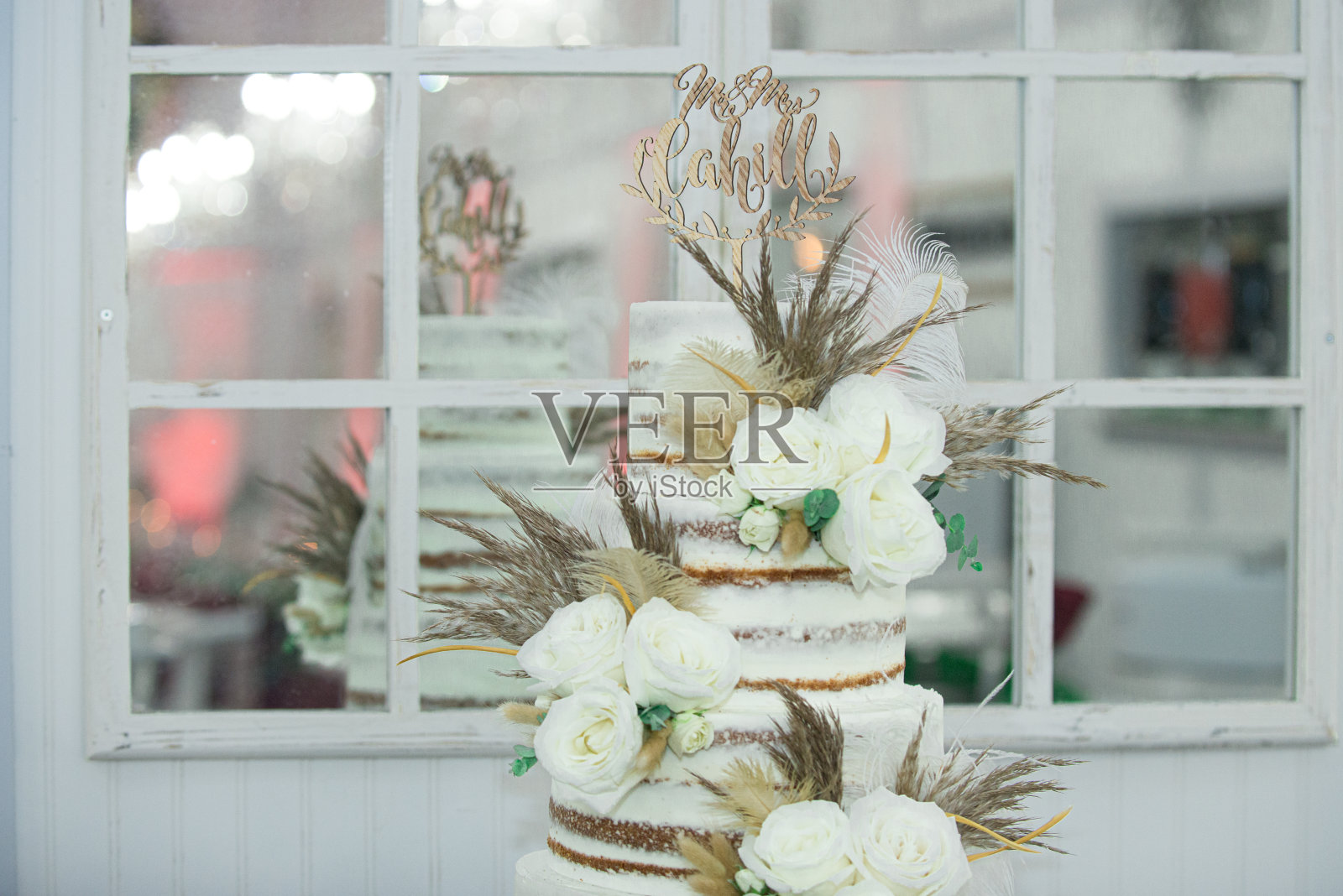三层裸婚蛋糕，装饰有白玫瑰和羽毛，窗户前有散景和反射照片摄影图片