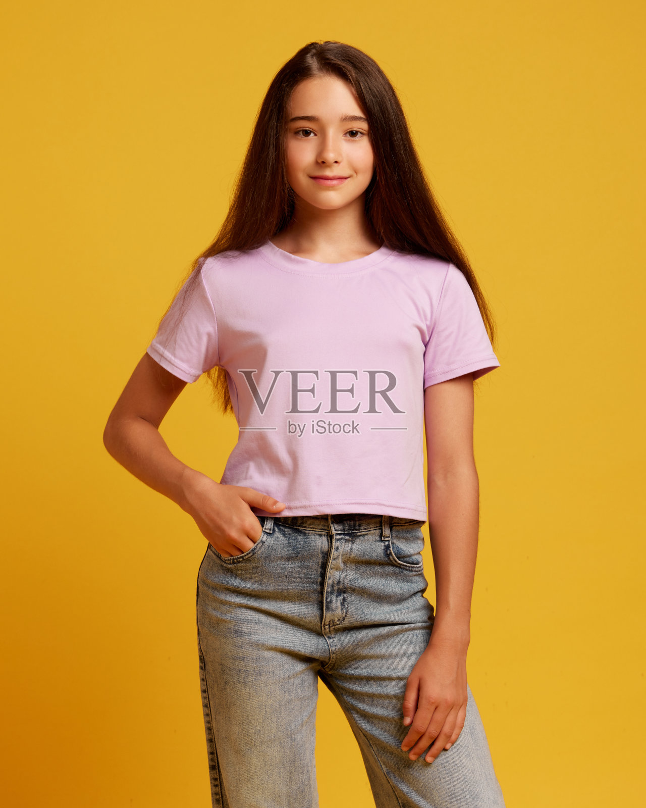 漂亮的十几岁女孩的肖像，棕色长发，穿着休闲牛仔裤和粉色t恤，站在黄色的工作室背景上带着微笑照片摄影图片