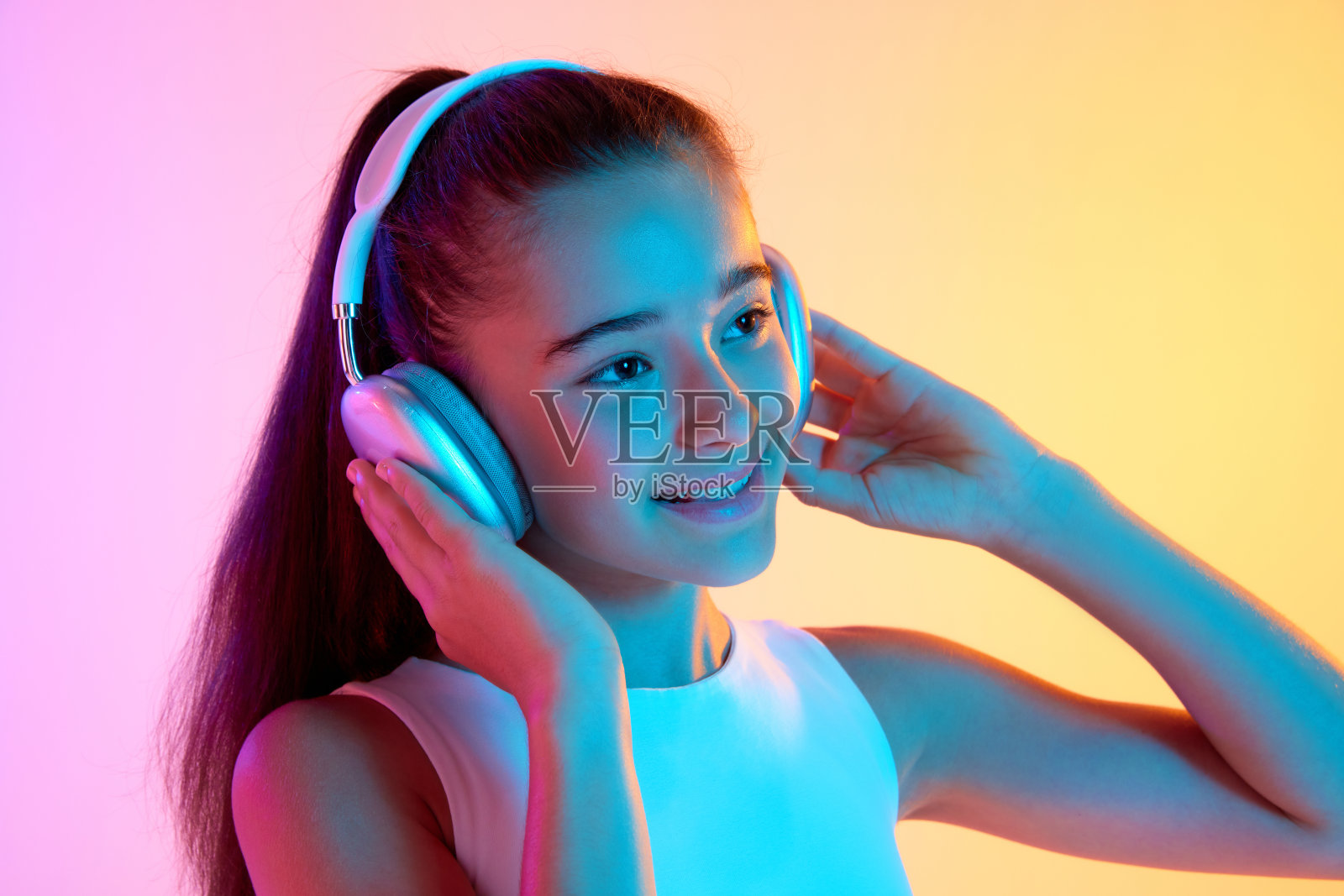 美丽的少女肖像，穿着白色上衣，戴着耳机听音乐，在霓虹灯的渐变粉黄色背景下微笑照片摄影图片