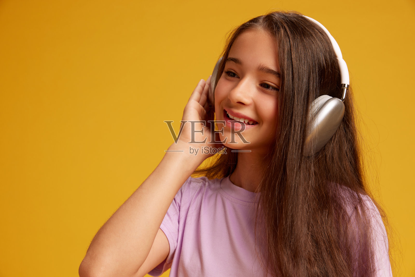 漂亮的十几岁女孩的肖像，棕色的长发，穿着粉红色的t恤，戴着耳机听音乐，在黄色的工作室背景下微笑照片摄影图片
