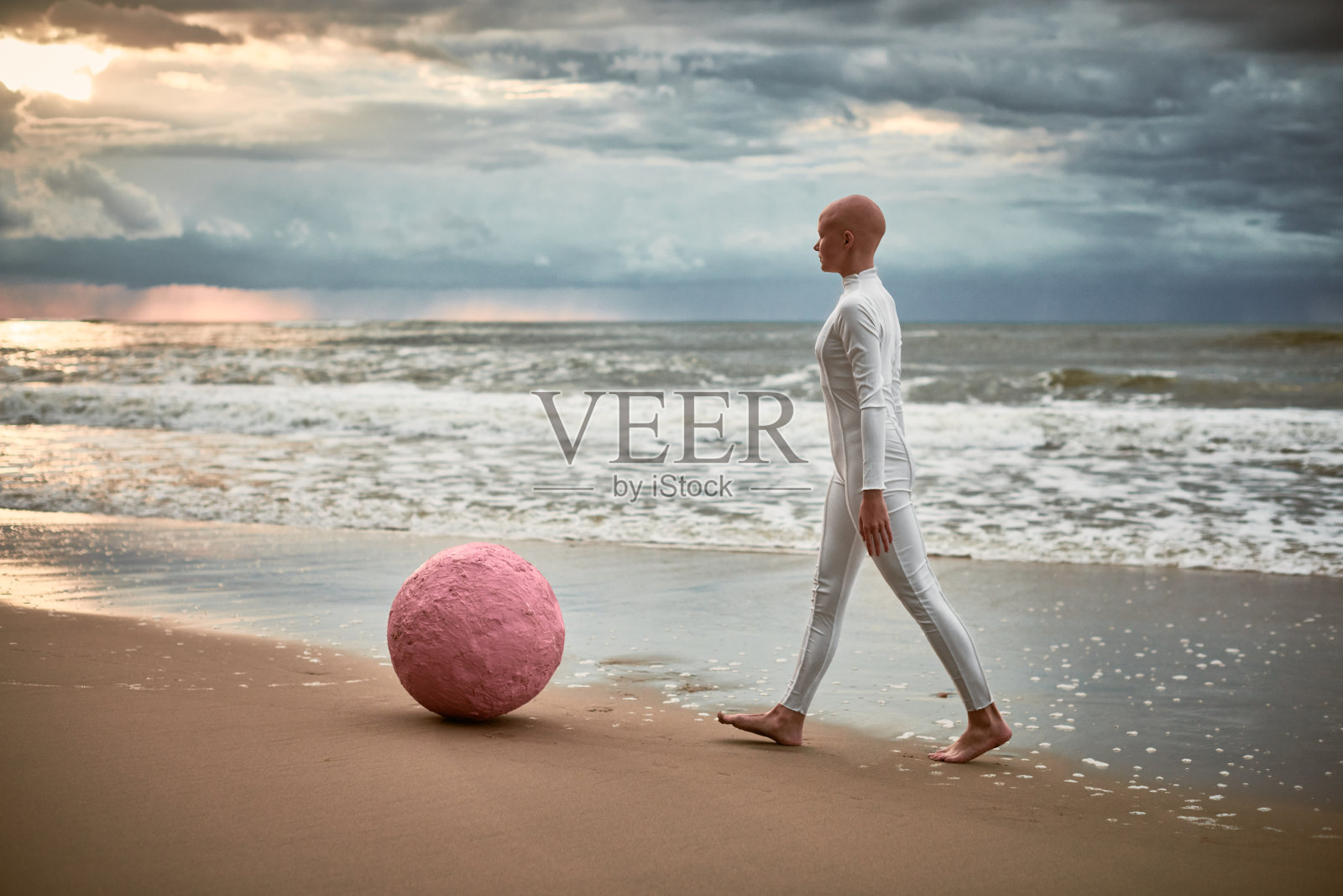 秃顶的表演女孩穿着未来派风格的西装，带着粉红色的球体沿着海岸散步照片摄影图片