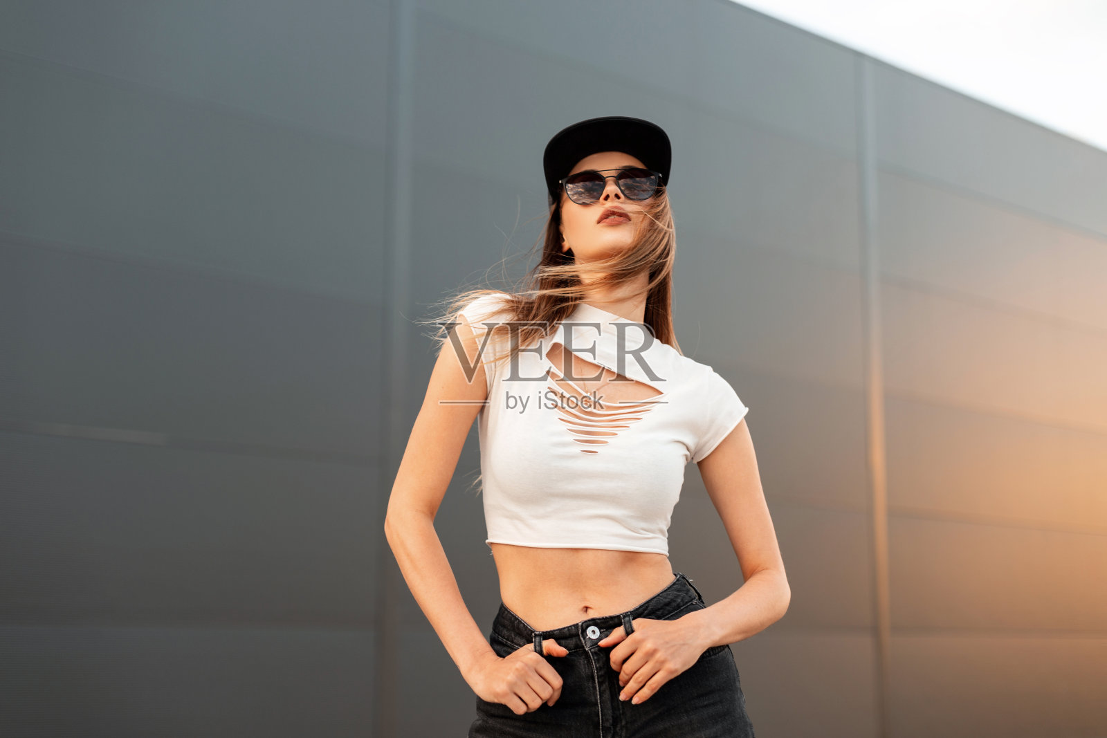 性感的时尚模特年轻女子戴着时尚的黑色太阳镜，穿着时髦的青年酷衣服，戴着美式黑色帽子，在橙色的夕阳下，在城市的复古墙旁摆姿势。休闲的夏季风格。照片摄影图片