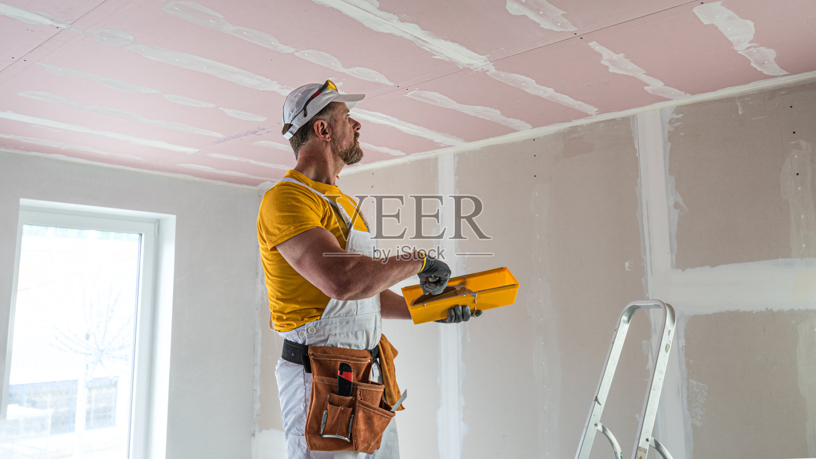 工人做了一个石膏板天花板。他在石膏干墙天花板接缝处粘胶带。红色石膏板提高天花板结构的防火性能。照片摄影图片