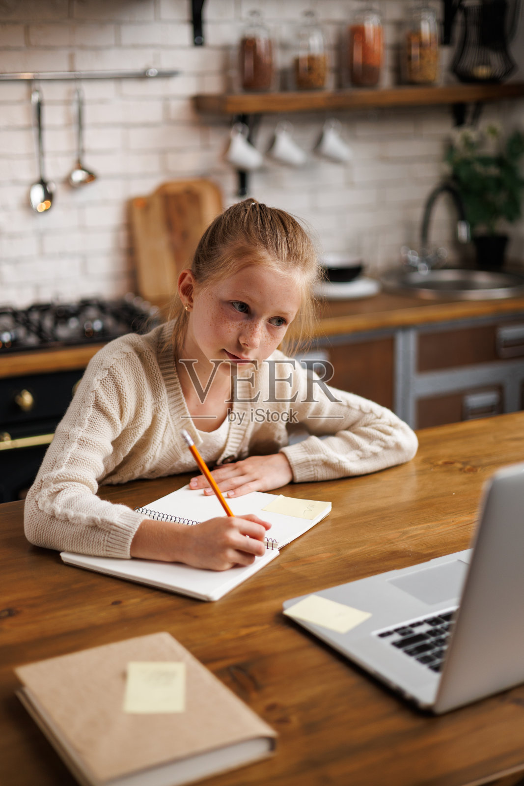 青春期前的女孩在家里的厨房里，在笔记本电脑和书本旁边的笔记本上写字照片摄影图片