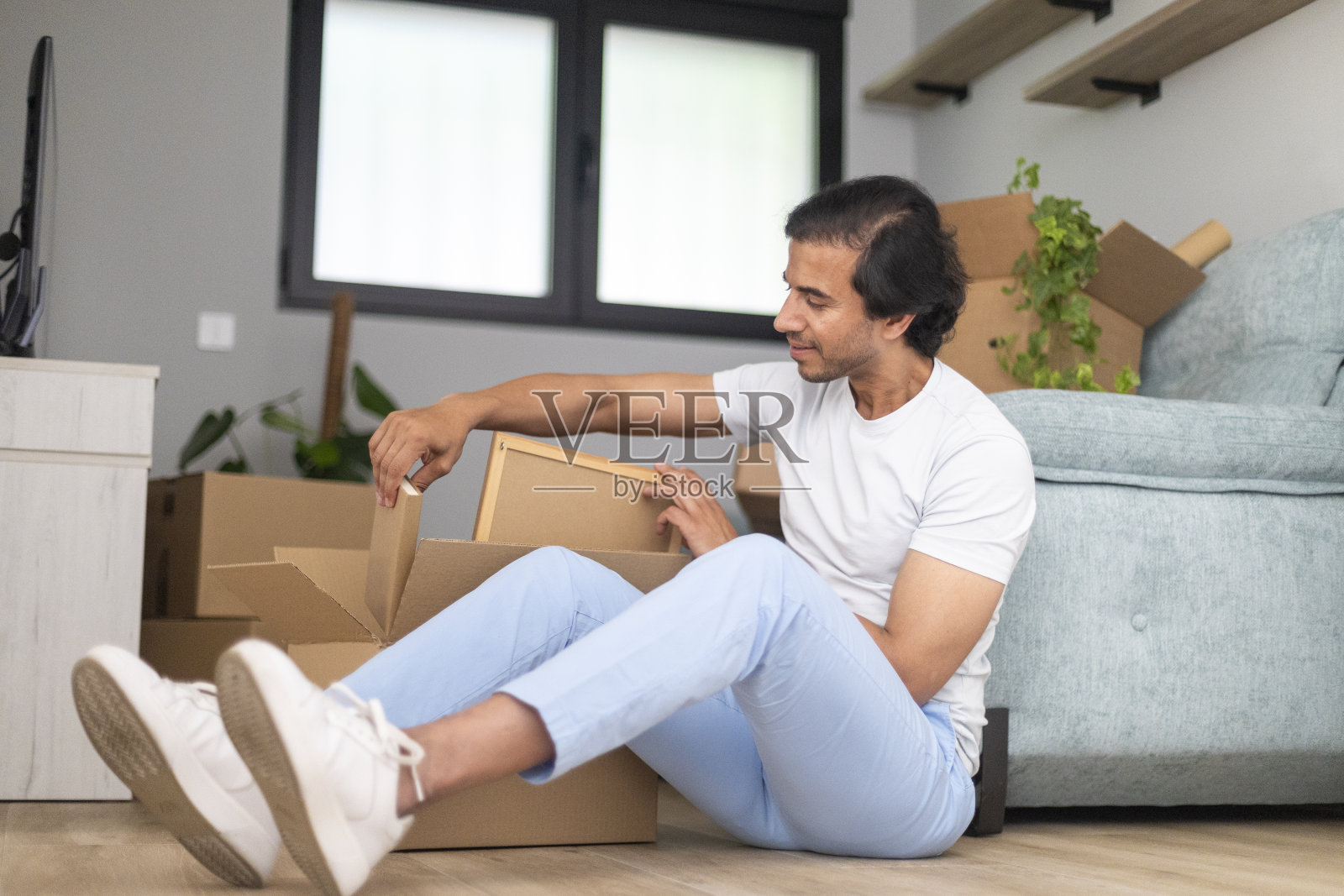 一名男子坐在新家的地板上，舒舒服服地打开箱子。照片摄影图片