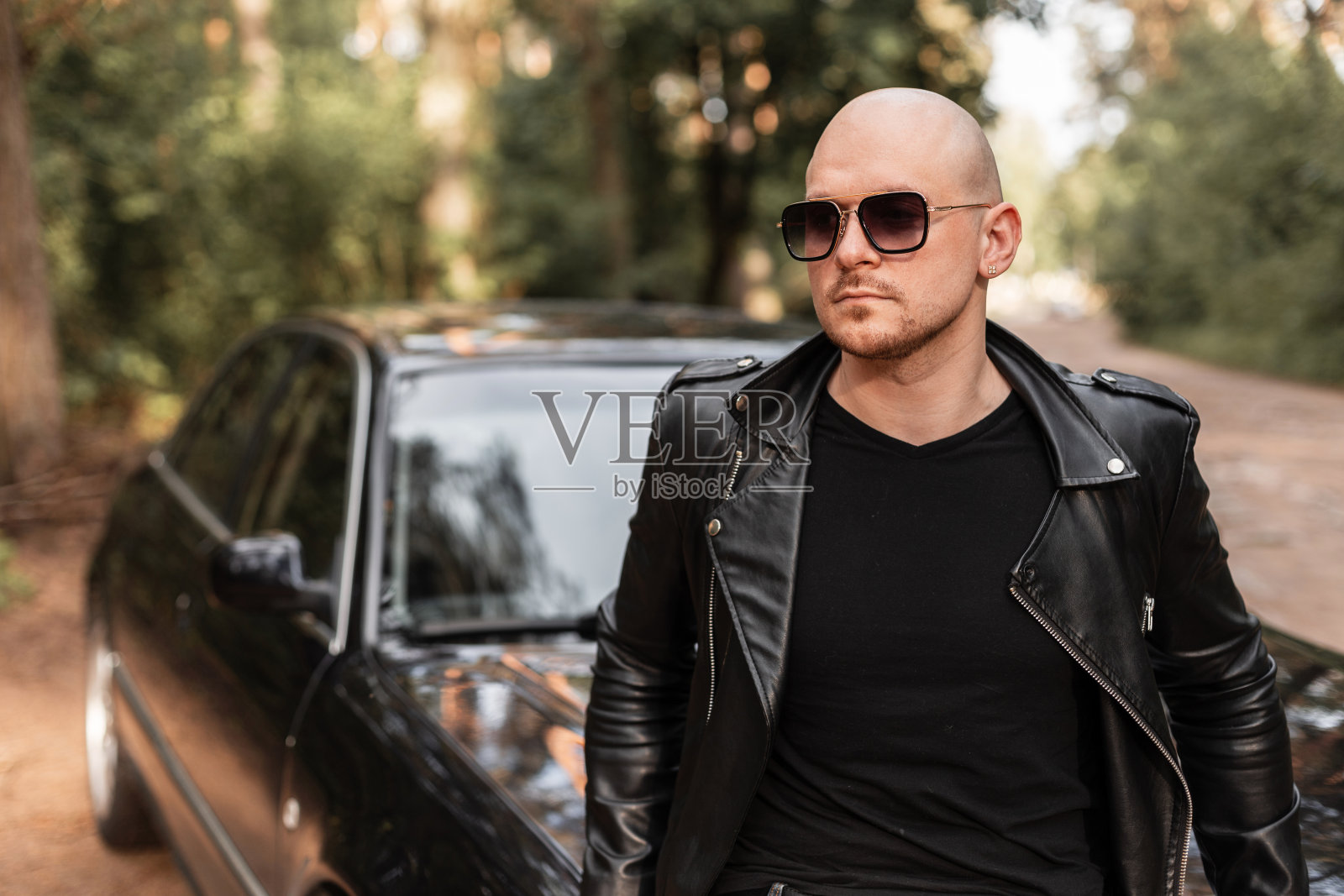 日落时分，穿着时尚皮夹克、戴着墨镜的帅哥站在一辆黑色轿车旁。野蛮的秃顶留着胡子的家伙在汽车长途旅行中休息。照片摄影图片