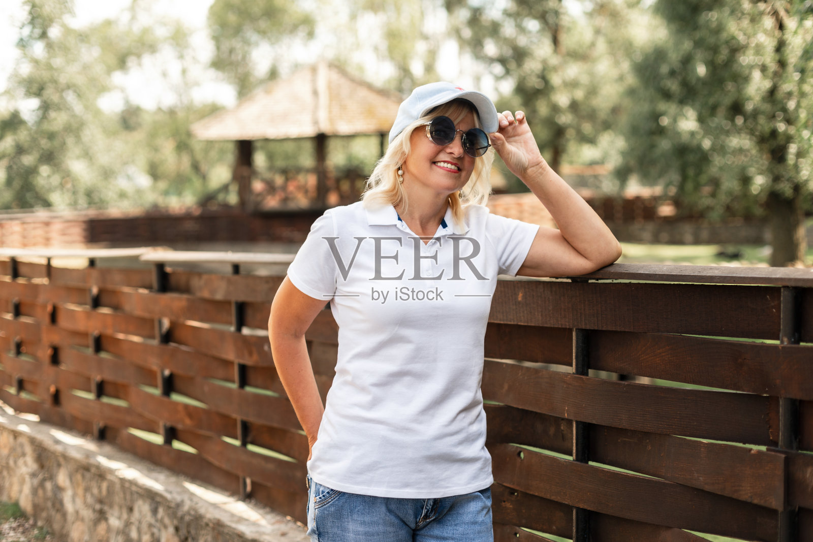 一位戴着墨镜、戴着帽子、穿着白色时尚polo t恤和蓝色牛仔裤的成年老妇人站在大自然的木栅栏旁照片摄影图片