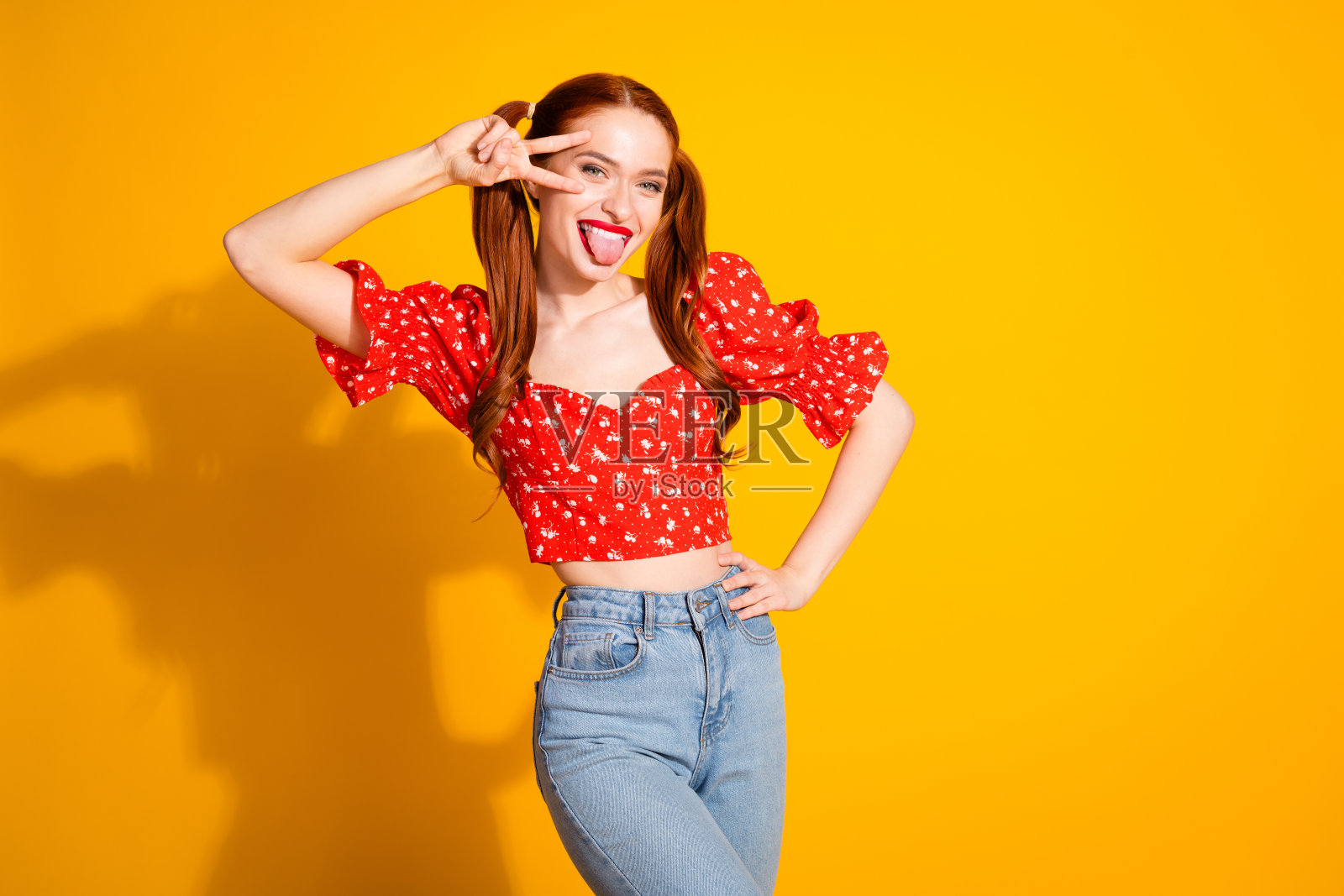 迷人的年轻女子的照片，露出v字的舌头，穿着时尚的红色衣服，孤立在黄色背景上照片摄影图片