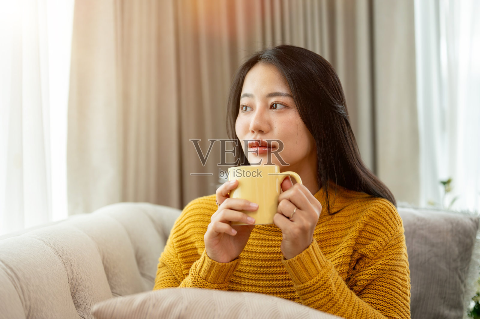 一个美丽的亚洲女人拿着咖啡杯，坐在沙发上，微笑着做着白日梦。照片摄影图片