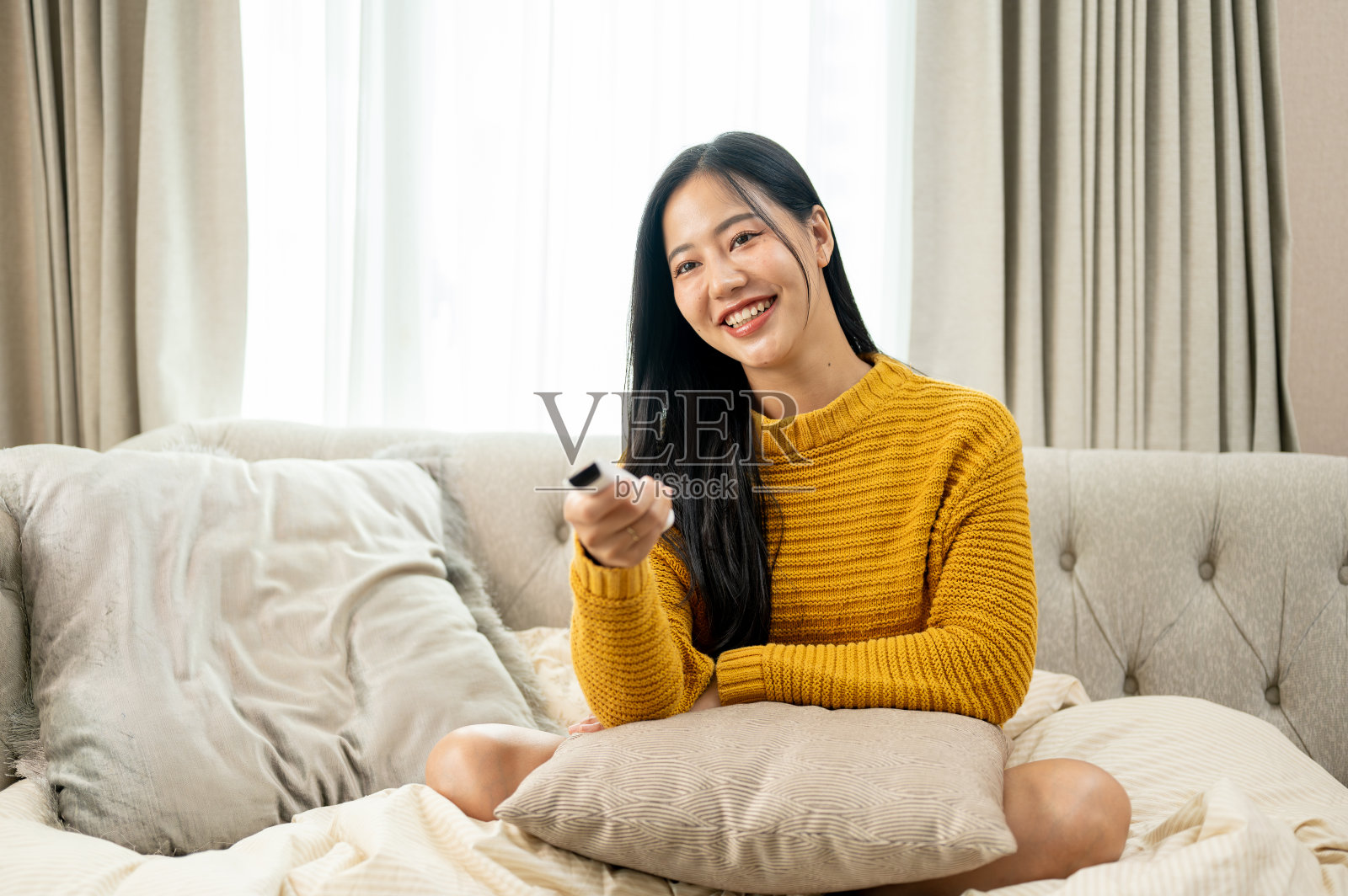 一位迷人的亚洲女性正拿着电视遥控器坐在客厅的沙发上看电视。照片摄影图片