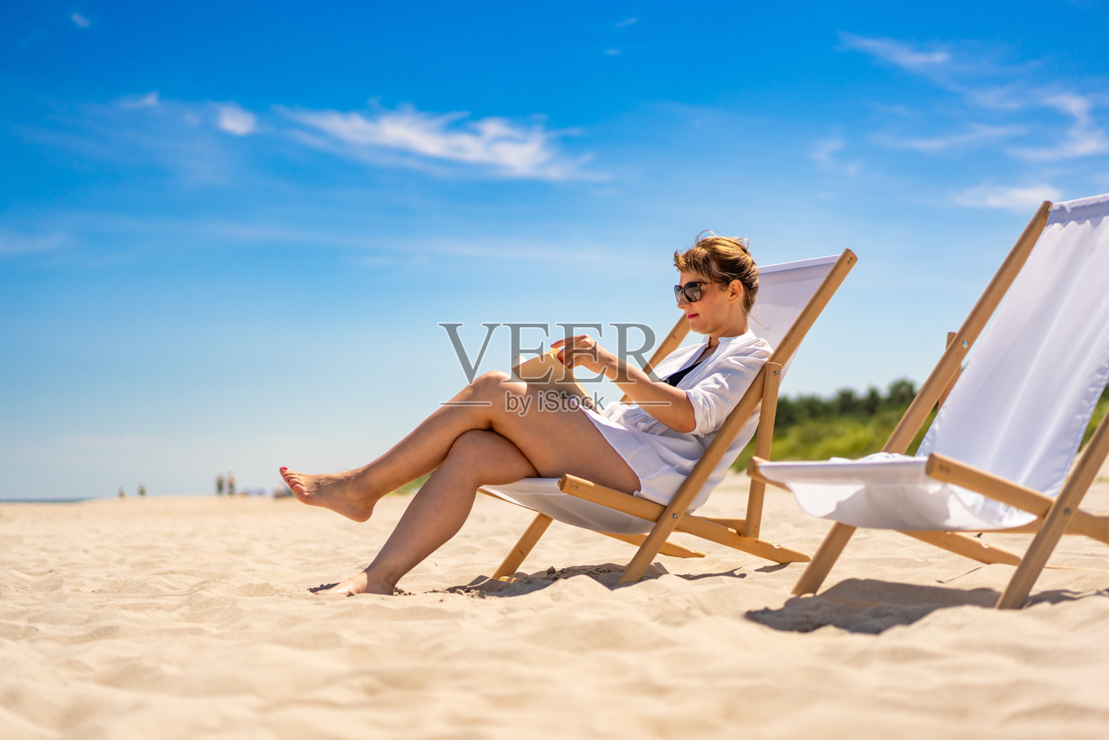 暑假在海滩上。在美丽的阳光明媚的日子里，穿着白衬衫的欧洲美女坐在沙滩上的躺椅上看书。在海滩上放松。侧视图照片摄影图片