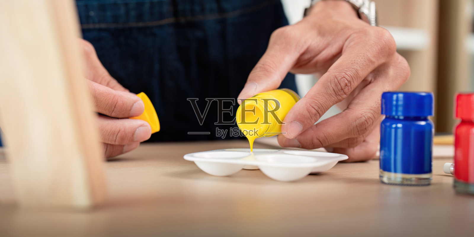 木桌上用蓝、红颜料瓶将黄色颜料倒入调色板的手特写照片摄影图片