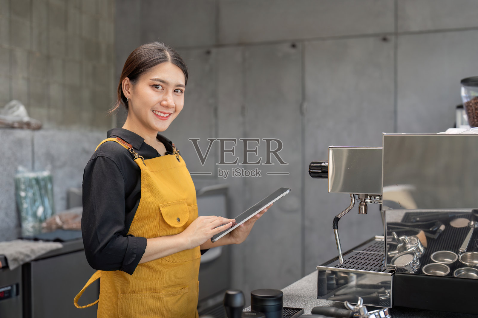 穿着黄围裙的年轻咖啡师在现代咖啡店使用平板电脑照片摄影图片