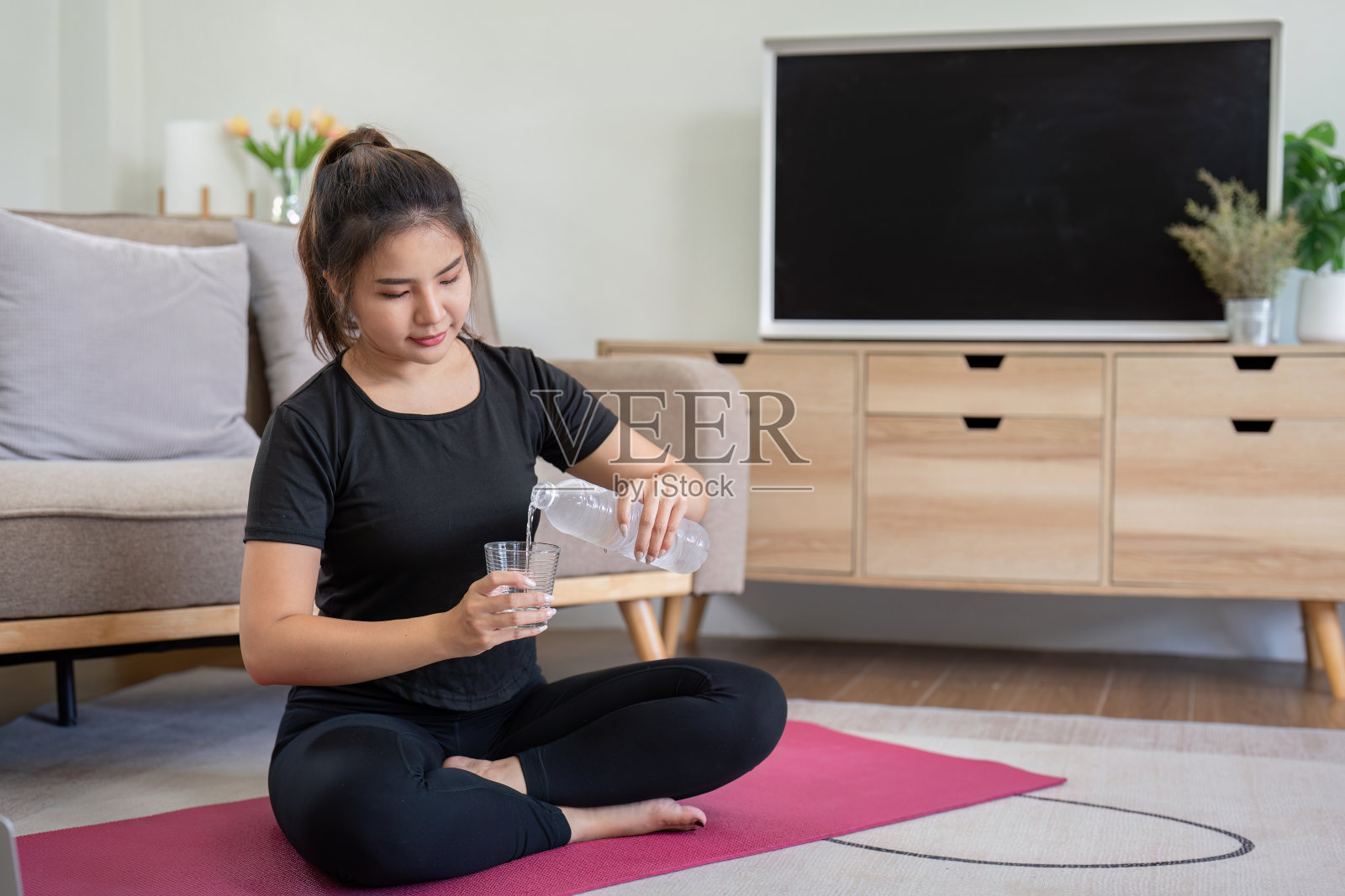 在舒适的客厅里，一位年轻女子一边在粉红色的垫子上往玻璃杯里倒水，一边练习瑜伽照片摄影图片